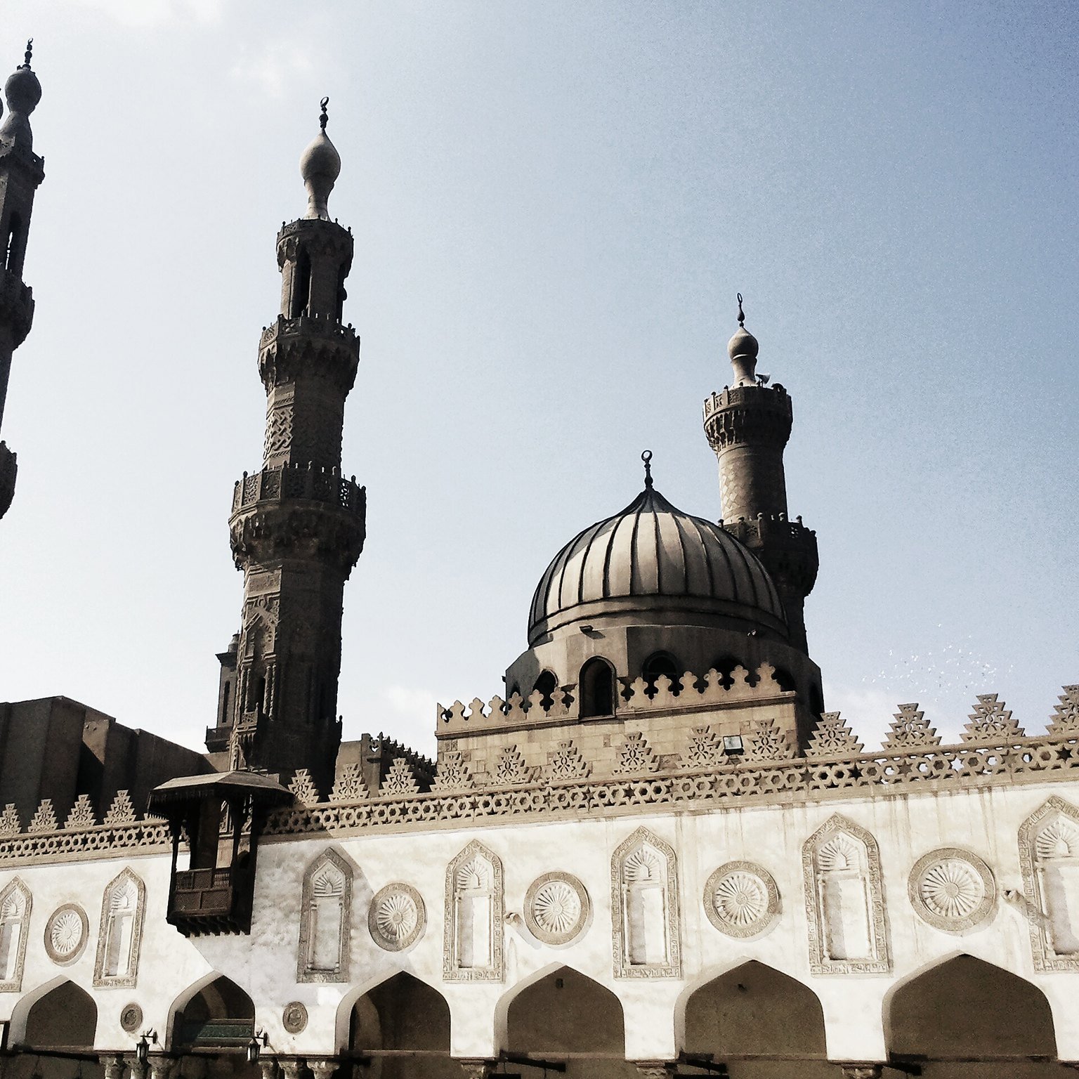 تظهر في الصورة مجموعة من المعالم المعمارية الإسلامية، حيث يتواجد مسجد كبير ذو قبتين ومئذنتين بارزتين. يبرز تصميم المسجد بتفاصيله المعمارية الفريدة، مثل النقوش والزخارف على الجدران. السماء خلف المبنى صافية، ما يضفي جواً من الهدوء على المشهد. تعكس الصورة الطابع التاريخي والثقافي للعمارة الإسلامية.