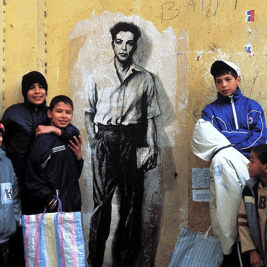 La imagen muestra a un grupo de niños posando al frente de una pared amarilla en la que hay un mural de un hombre en blanco y negro. Los niños parecen estar disfrutando del momento y están vestidos con ropa de abrigo. Algunos de ellos sostienen bolsas, posiblemente de tela. La escena transmite una sensación de comunidad y alegría, en contraste con el fondo urbano y desgastado de la pared.