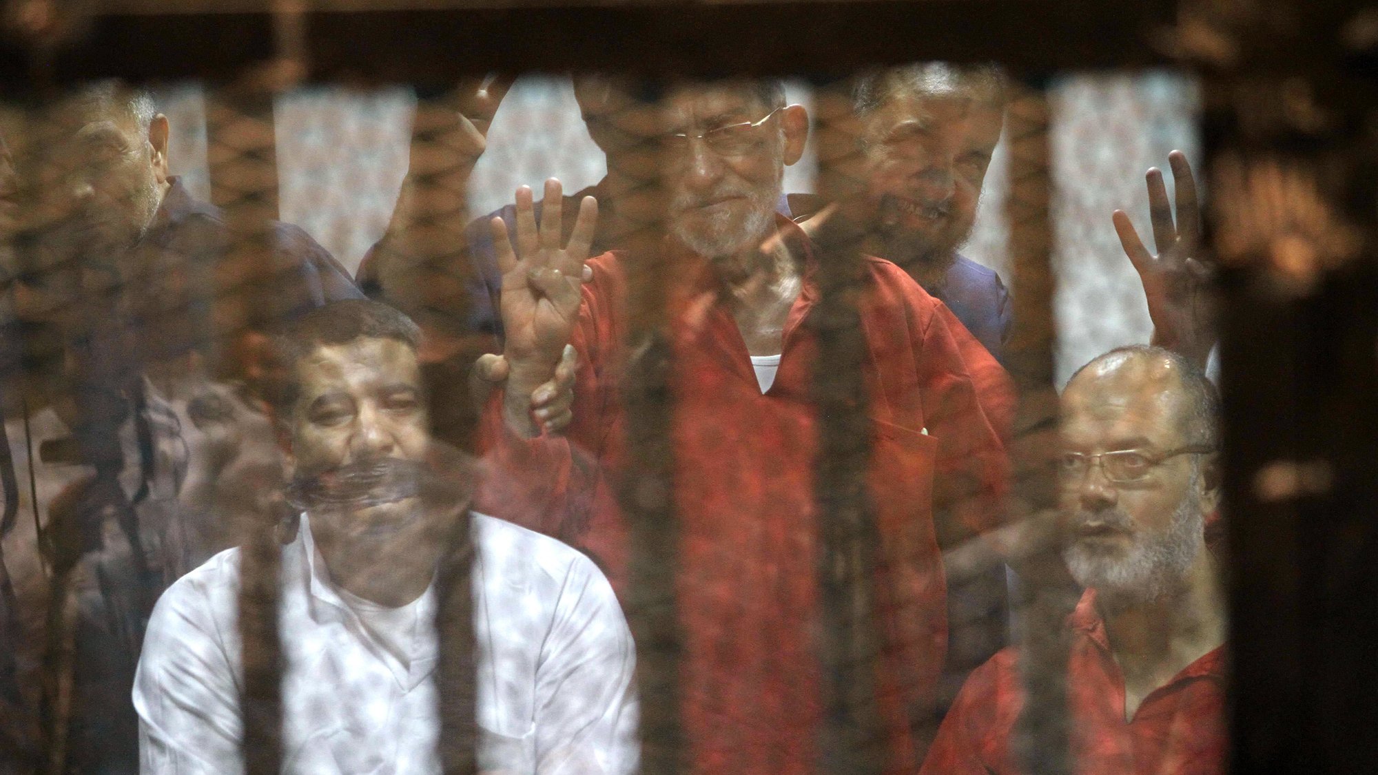 L'image montre un groupe d'hommes derrière une grille ou un panneau de verre. Ils semblent être dans un contexte de détention ou de procès, avec des expressions variées sur leurs visages. Certains lèvent les mains, suggérant un geste de protestation ou de solidarité. L'ambiance générale est sombre, évoquant des thèmes de justice et de droits humains.