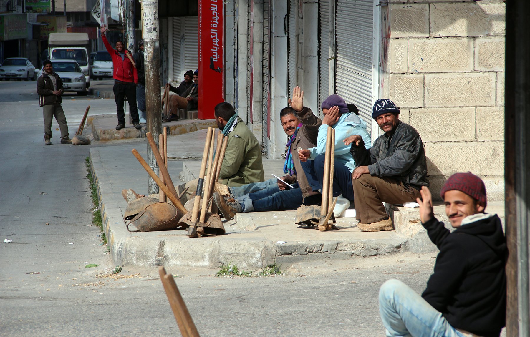 L'image montre un groupe d'hommes assis sur le bord de la route, apparemment en attente. Ils semblent discuter et certains saluent la caméra. L'environnement a l'air urbain, avec des bâtiments en arrière-plan et une rue. Des outils de travail sont visibles, suggérant qu'ils pourraient être des travailleurs. L'atmosphère semble décontractée malgré le cadre urbain.