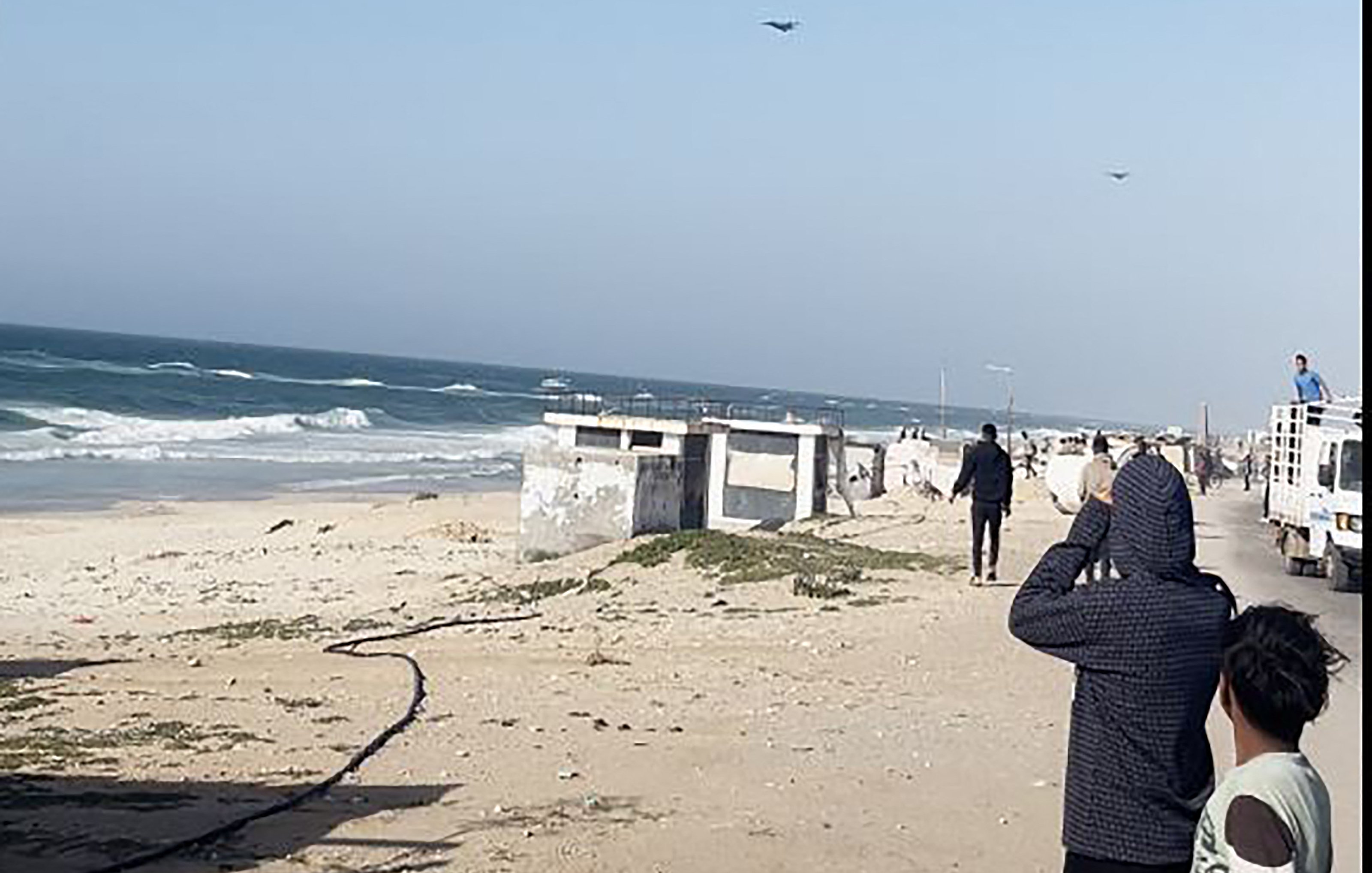 L'image montre une scène côtière avec des vagues de mer s'écrasant sur le rivage. Au premier plan, plusieurs personnes regardent vers l'horizon, peut-être fascinées par quelque chose dans le ciel. On peut également voir des véhicules stationnés sur le côté. L'ambiance semble calme, avec un ciel dégagé et une légère brise. La plage est principalement sablonneuse, et quelques éléments tels que des algues sont visibles sur le sol.