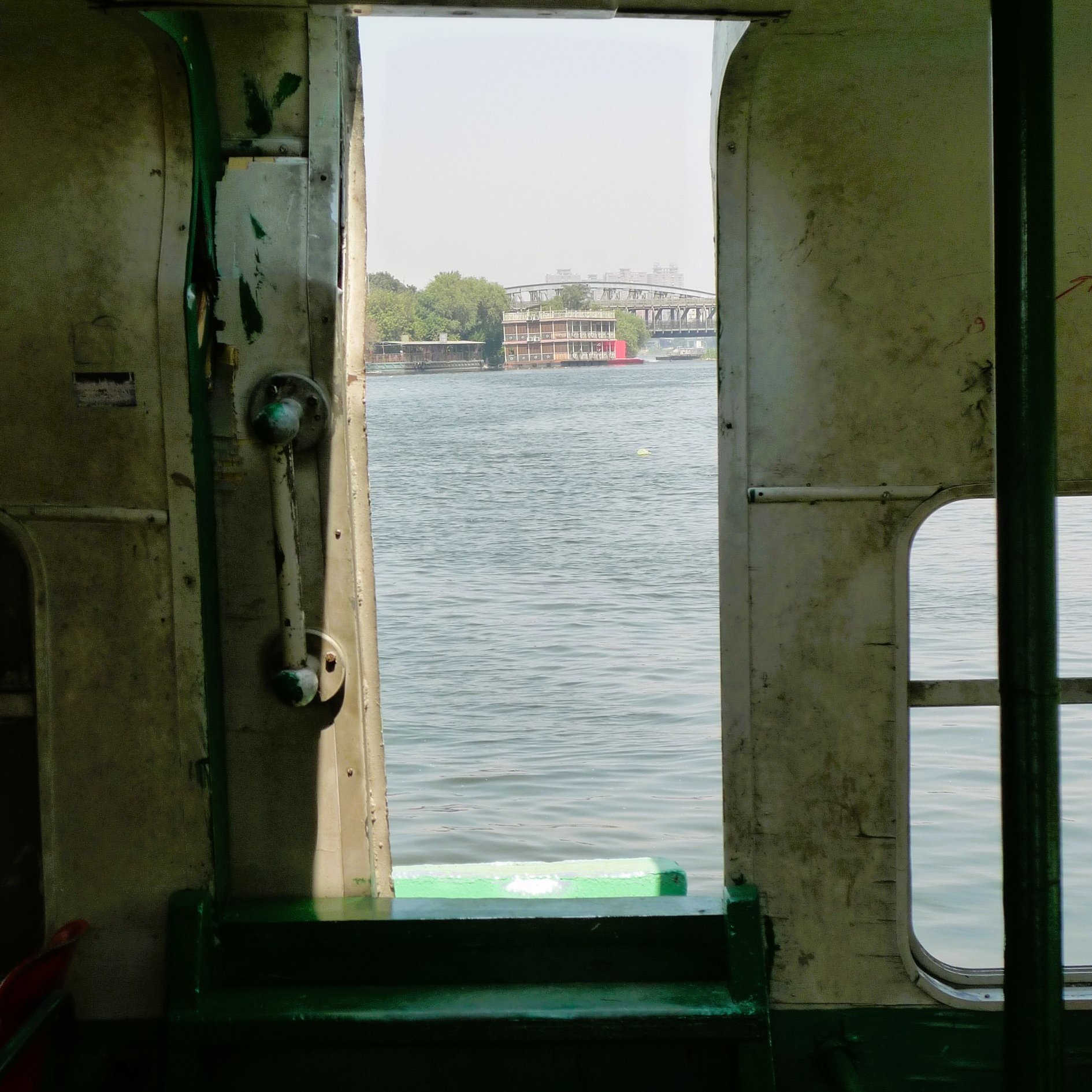 L'immagine mostra l'interno di un'imbarcazione, con una porta aperta che si affaccia su un corpo d'acqua. La vista rivela un paesaggio sereno, con un riflesso dell'acqua e vegetazione visibile in lontananza. La struttura della barca appare semplice, con pareti in metallo e sedili visibili all'interno. La luce naturale entra attraverso l'apertura, creando un'atmosfera tranquilla.
