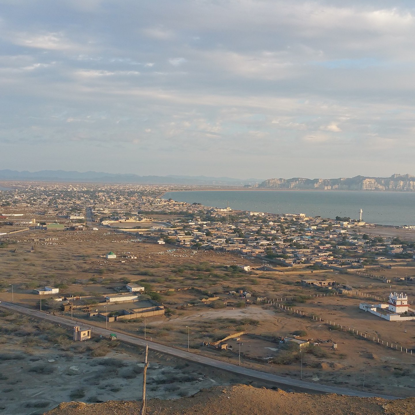 L'image montre un paysage côtier avec une vue panoramique. On peut voir une ville située au bord de la mer, entourée de collines et de montagnes à l'horizon. La mer est calme, et des structures côtières, comme des docks ou des bateaux, sont visibles. Le ciel est partiellement nuageux, ce qui donne une belle lumière à la scène. On observe également des zones désertiques ou semi-désertiques, avec des routes qui serpentent à travers le paysage.