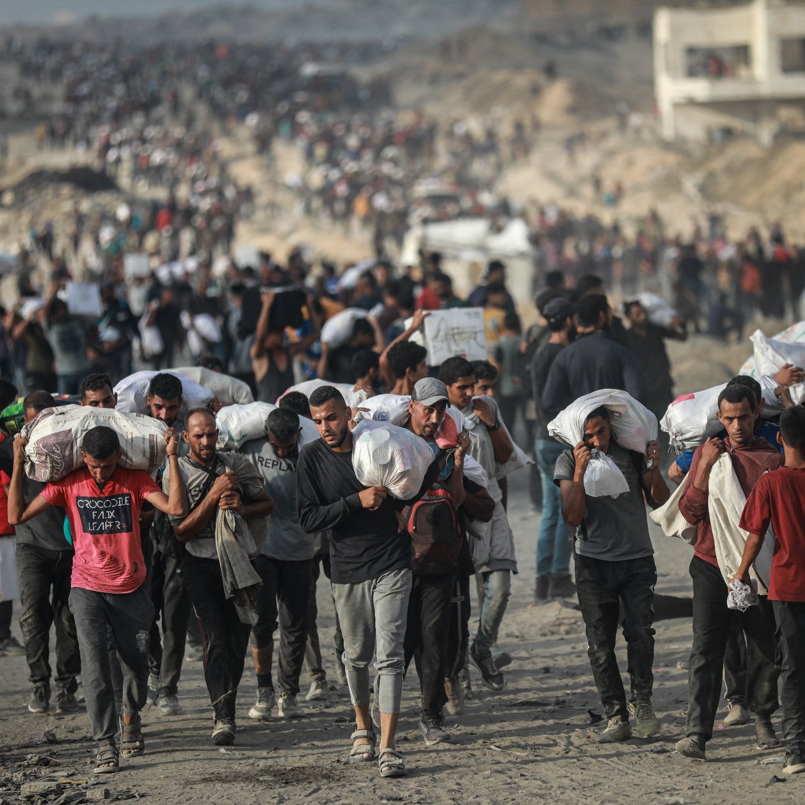 Gruppo di persone che trasportano sacchi su un terreno polveroso, in un contesto affollato.