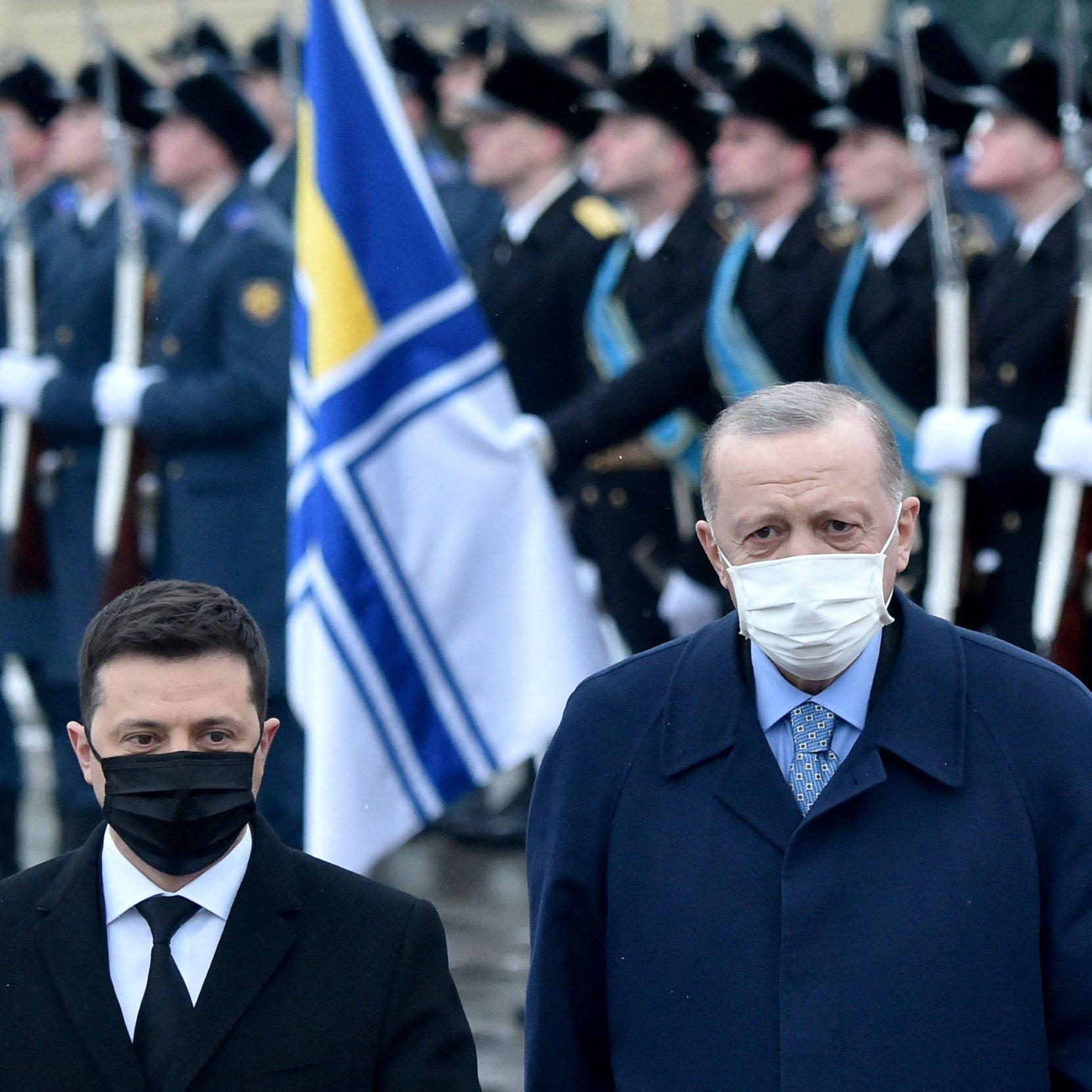 The image shows two men standing side by side in a formal setting, likely during a military ceremony or event. One man is wearing a black face mask and dark suit, and the other is in a blue coat and also wearing a mask. In the background, there is a formation of soldiers in uniform holding rifles, and a flag is prominently displayed, suggesting a significant occasion. The overall atmosphere appears solemn and official.