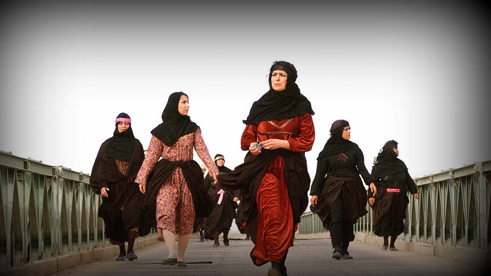 L'image montre un groupe de femmes marchant sur un pont. Elles portent des vêtements traditionnels, principalement des robes longues et des foulards. La scène semble être en extérieur, avec un ciel clair en arrière-plan. Les femmes ont des expressions déterminées et se déplacent ensemble, ce qui dégage une impression d'unité et de force.