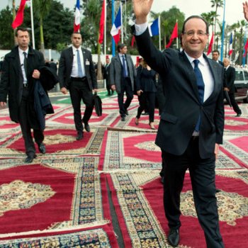 L'image montre deux hommes marchant sur un tapis richement décoré. L'un d'eux, vêtu d'un costume sombre et souriant, fait un geste de la main, tandis que l'autre, également en costume, marche à ses côtés. En arrière-plan, on peut voir des drapeaux de différentes nations, ce qui suggère un événement international ou officiel. L'atmosphère semble conviviale et solennelle à la fois.
