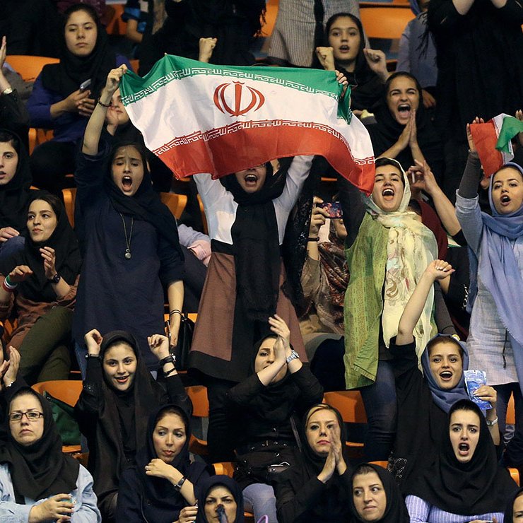 L'image montre un groupe de femmes dans une arène, probablement lors d'un événement sportif. Elles portent des vêtements traditionnels et modernes, et certaines d'entre elles agitent un drapeau iranien, exprimant leur soutien et leur enthousiasme. Les visages sont joyeux et animés, et l'atmosphère semble festive. Ce moment illustre la participation et la passion des femmes pour le sport en Iran.