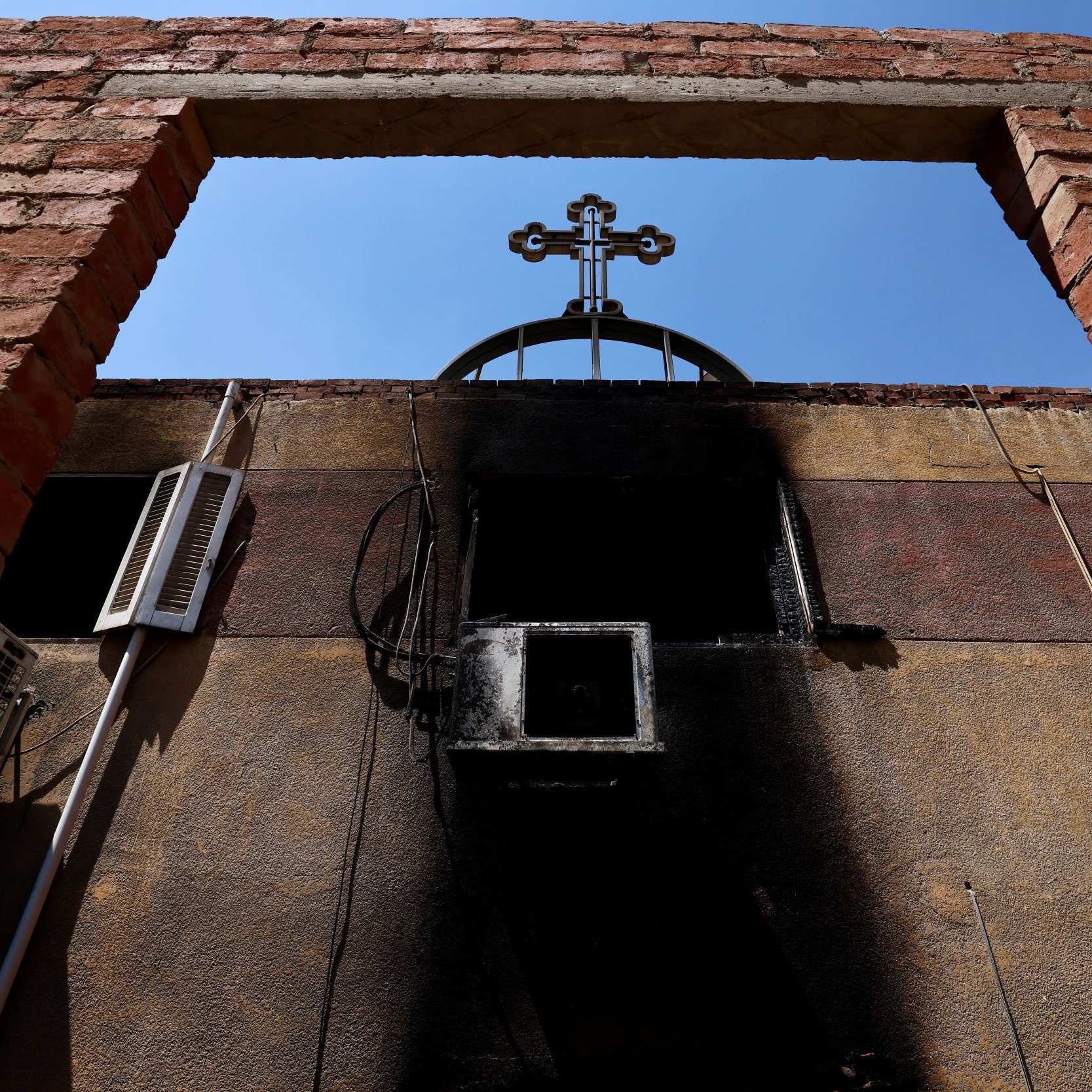 الصورة تظهر إطارًا من الطوب الأحمر مع فتحة في الجزء العلوي، ومن خلال هذه الفتحة يمكن رؤية صليب مرفوع في السماء. الجدران المحيطة تبدو متضررة، حيث يظهر عليها أثر السواد، مما يشير إلى وجود حريق أو تلف. السماء في الخلفية زرقاء وصافية، مما يعكس تناقضًا مع الأجواء المحيطة.