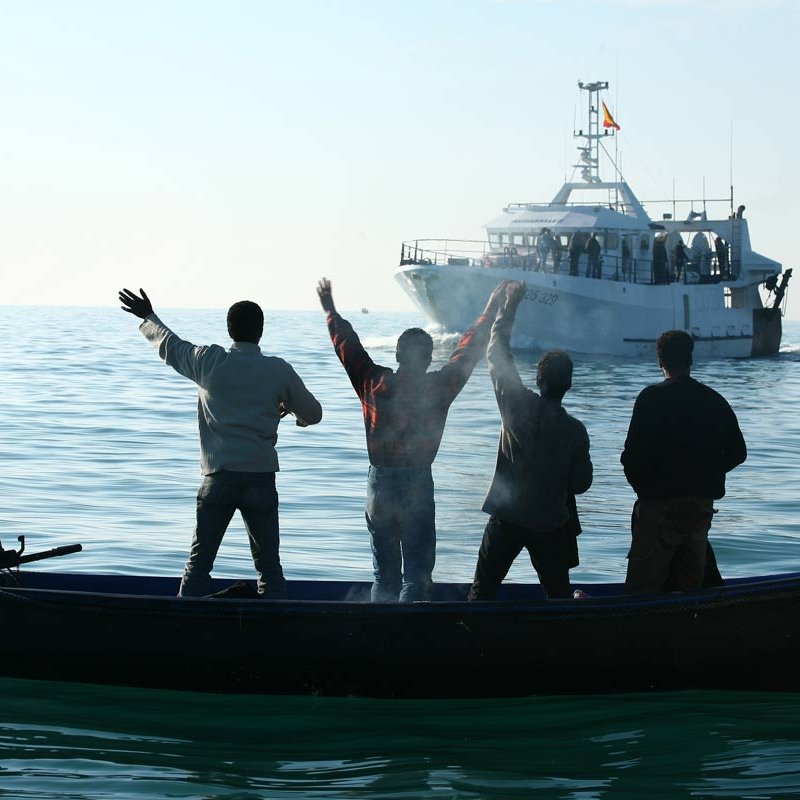 تظهر في الصورة مجموعة من الأشخاص يقفون في قارب صغير في المياه الهادئة، ويبدو أنهم يلوحون بأيديهم نحو قارب أكبر يقترب منهم. المشهد يعكس نشاطًا بحريًا، حيث الأجواء هادئة وتظهر السماء والماء بلونٍ صافٍ. الأشخاص يظهرون من الخلف، مما يركز الانتباه على الإيماءات التي يقومون بها.