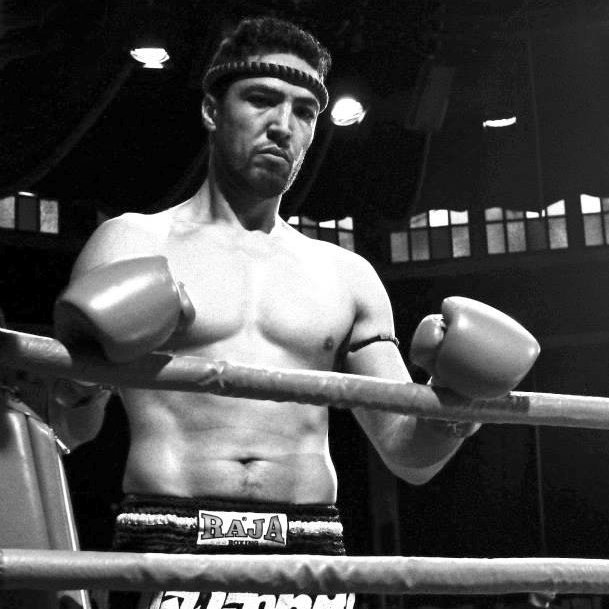 الصورة تظهر الملاكم في حلبة الملاكمة، حيث يبدو عابسًا وجادًا. هو يرتدي حزامًا أو شورت رياضي مكتوب عليه "RAJA" وتظهر على يديه قفازات الملاكمة. تعكس الإضاءة ونمط الصورة باللونين الأبيض والأسود أجواء المنافسة والقتال.