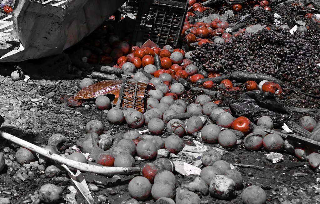 تظهر الصورة مجموعة من الفواكه المتناثرة على الأرض. الألوان الرئيسية هي تدرجات رمادية، مع تواجد بعض الفواكه باللون الأحمر، مثل الطماطم. هناك أيضًا بعض السلال المستخدمة لتخزين الفواكه، وبعض الفواكه الأخرى التي تبدو متروكة ومهملة. تبدو البيئة المحيطة غير مرتبة، مما يشير إلى حالة من الفوضى أو الإهمال.