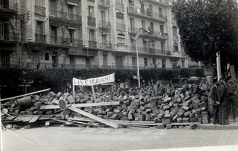 L'image montre une scène historique avec un amas de briques et de débris, suggérant une barricade ou un barrissement. Au premier plan, on peut voir des personnes rassemblées, probablement des manifestants ou des membres d'une foule, observant la scène. Un panneau est visible, ce qui pourrait indiquer le message ou le motif de la manifestation. À l'arrière-plan, des bâtiments avec des balcons sont présents, typiques d'une architecture urbaine. L'atmosphère semble tendue, reflétant un moment de contestation ou de protestation.