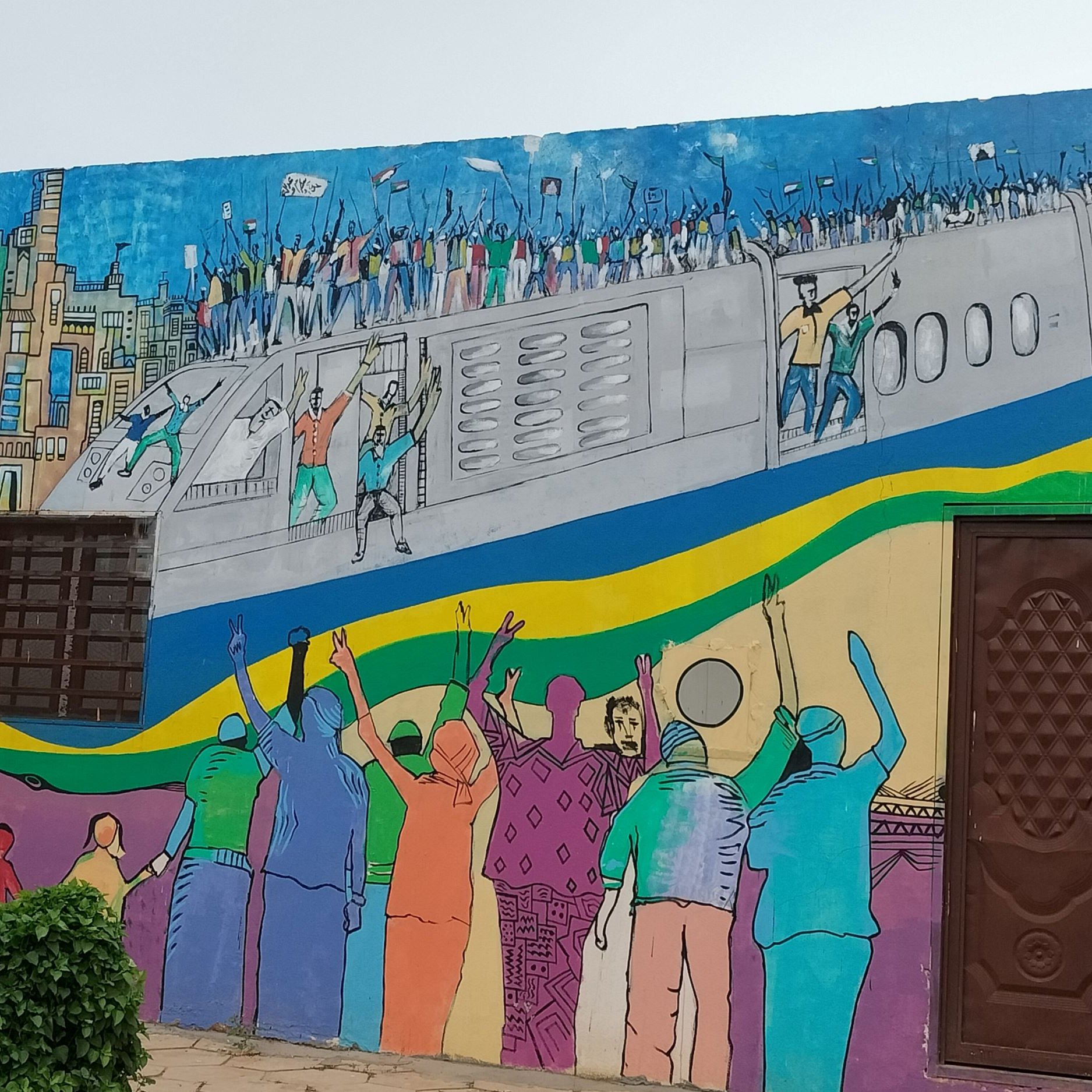 L'immagine rappresenta un murale colorato su un muro. La scena ritrae una folla di persone che sembra celebrare o protestare, con alcune figure che sollevano le braccia in segno di gioia o di richiesta. Sullo sfondo, c'è un aereo che si staglia sopra una città stilizzata, caratterizzata da edifici e una vivace palette di colori. I colori utilizzati nel murale sono brillanti e vivaci, riflettendo un senso di energia e movimento. La composizione comunica un forte messaggio di unità e di movimento collettivo.