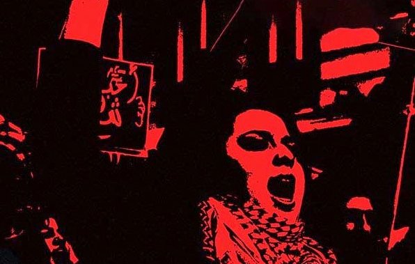 L'image présente une scène expressive et énergique, probablement de manifestation ou de rassemblement. On y voit une personne au premier plan qui lève son bras, suggérant un appel à l'action ou une déclaration de protestation. L'arrière-plan est dominé par des couleurs sombres, ce qui intensifie le sentiment de détermination. Des affiches ou des slogans en arabe sont visibles, renforçant l'idée d'un message collectif. L'atmosphère évoque un fort engagement et une passion pour une cause.