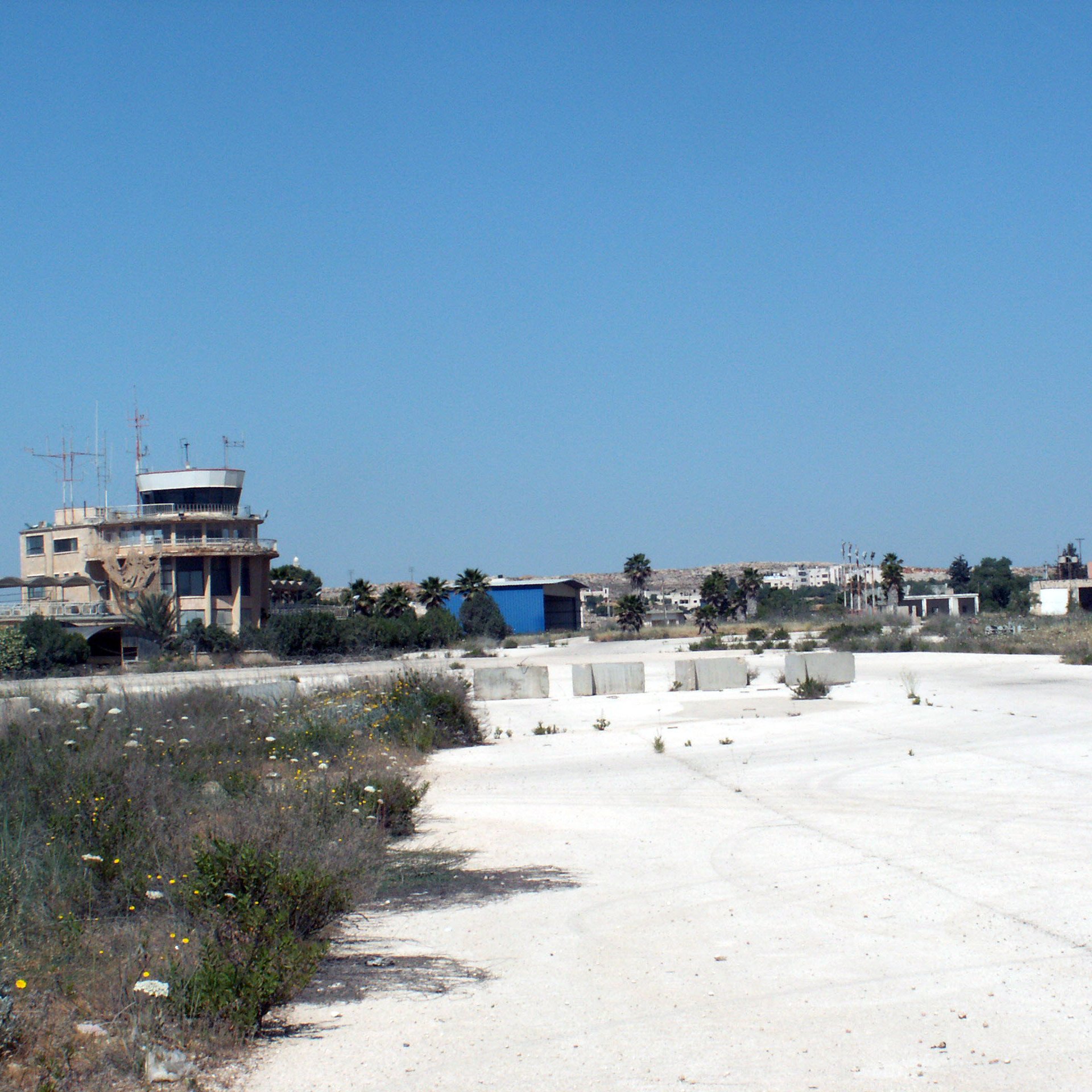 The image depicts an abandoned or derelict area, likely an old airstrip or airport. In the background, there is a control tower or building, which appears to be in disrepair. Surrounding the area are sparse patches of vegetation, including some wildflowers, and the ground is mostly composed of white gravel or concrete. The sky is clear and blue, indicating a bright, sunny day, and the overall scene conveys an atmosphere of neglect and desolation.