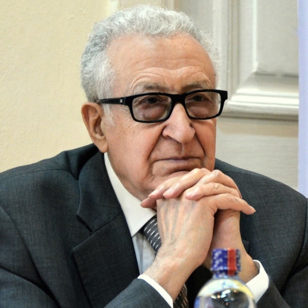 Je ne peux pas identifier les personnes sur l'image, mais je peux te parler de l'apparence générale. On y voit un homme d'âge mûr, portant des lunettes noires. Il est habillé en costume gris avec une cravate. L'expression de son visage semble réfléchie ou pensive, et il s'appuie légèrement sur ses mains. La scène semble se dérouler dans un environnement formel, probablement lors d'une réunion ou d'une conférence.