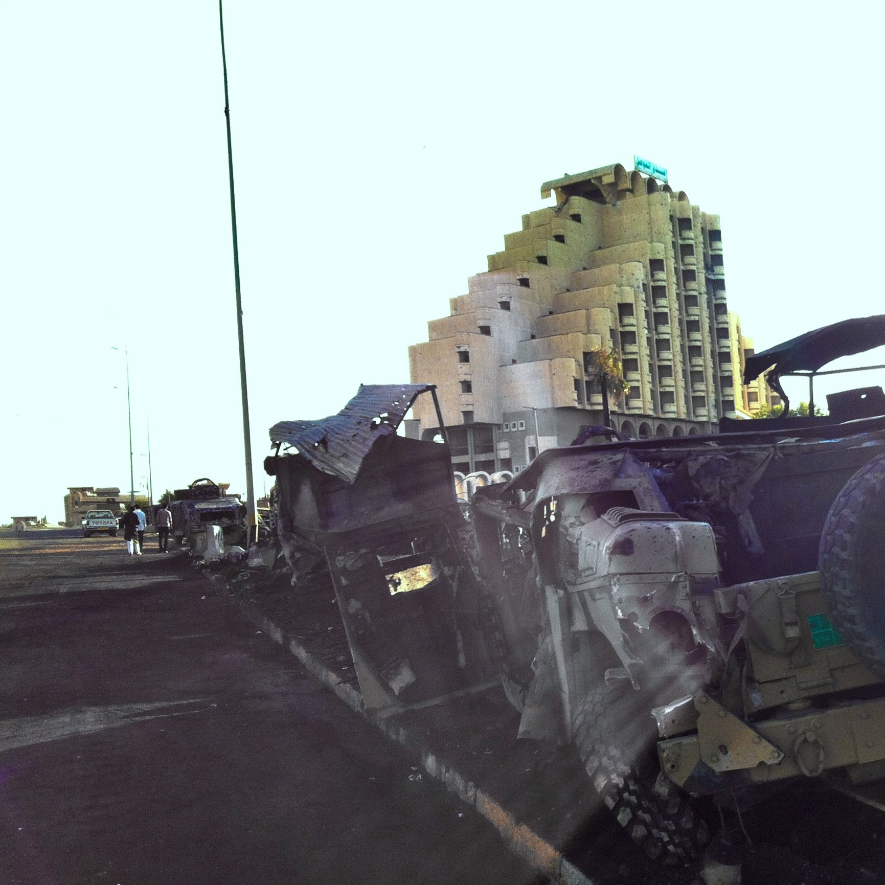 The image depicts a scene with damaged military vehicles, likely military trucks, that appear to be abandoned or destroyed along a road. In the background, there is a large, partially constructed building with a distinctive pyramid-like shape. The setting suggests a location that has experienced conflict or destruction, and the lighting gives the scene a bright, almost surreal quality. There are a few individuals visible in the background, indicating some activity in the area. Overall, the image conveys a sense of desolation and the remnants of past turmoil.