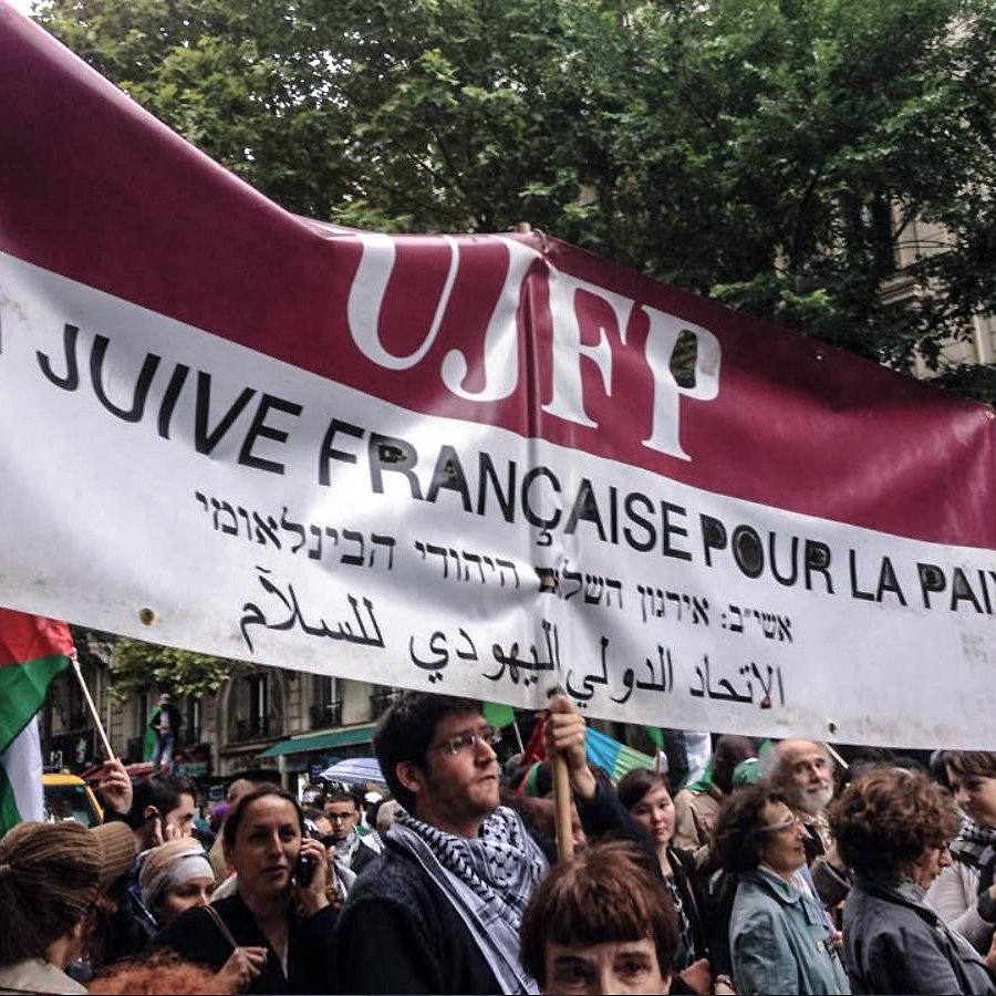 الصورة تظهر تجمعًا حاشدًا للأشخاص في مسيرة، حيث يرفع المشاركون لافتة كبيرة. اللافتة مكتوبة باللغات الفرنسية والعبرية والعربية، وتعبّر عن التضامن من أجل السلام. يبدو أن هناك تنوعًا في المشاركين، حيث يرتدي البعض الزي التقليدي أو يحملون أعلامًا مختلفة. الأجواء في الصورة تعكس روح النضال والمطالبة بحقوق معينة.