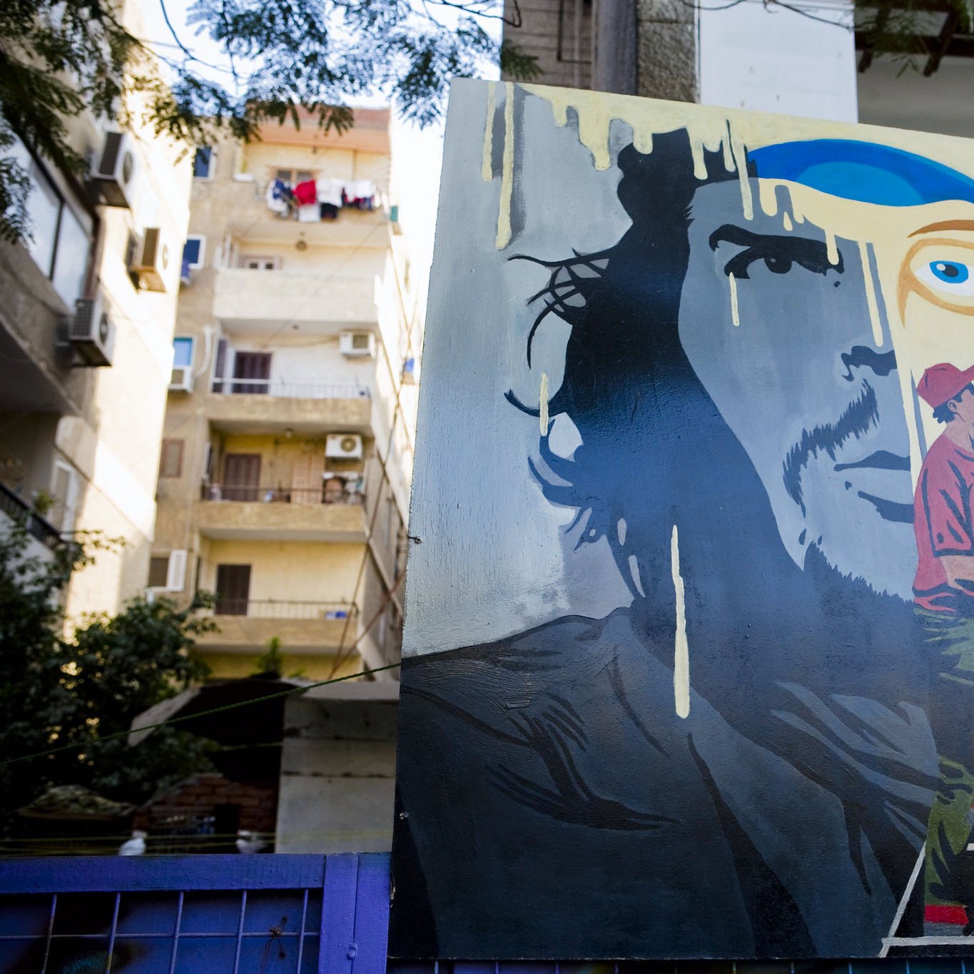 L'immagine mostra un murale artistico su un muro esterno. Il dipinto raffigura il volto di un uomo con un'espressione intensa, accanto a un artista in azione, intento a dipingere. Le tonalità utilizzate sono vivaci, con predominanza di blu e verde, creando un contrasto interessante. Sullo sfondo, si intravedono elementi architettonici e tende, suggerendo un ambiente urbano. Questo murale esprime una fusione tra arte e quotidianità.