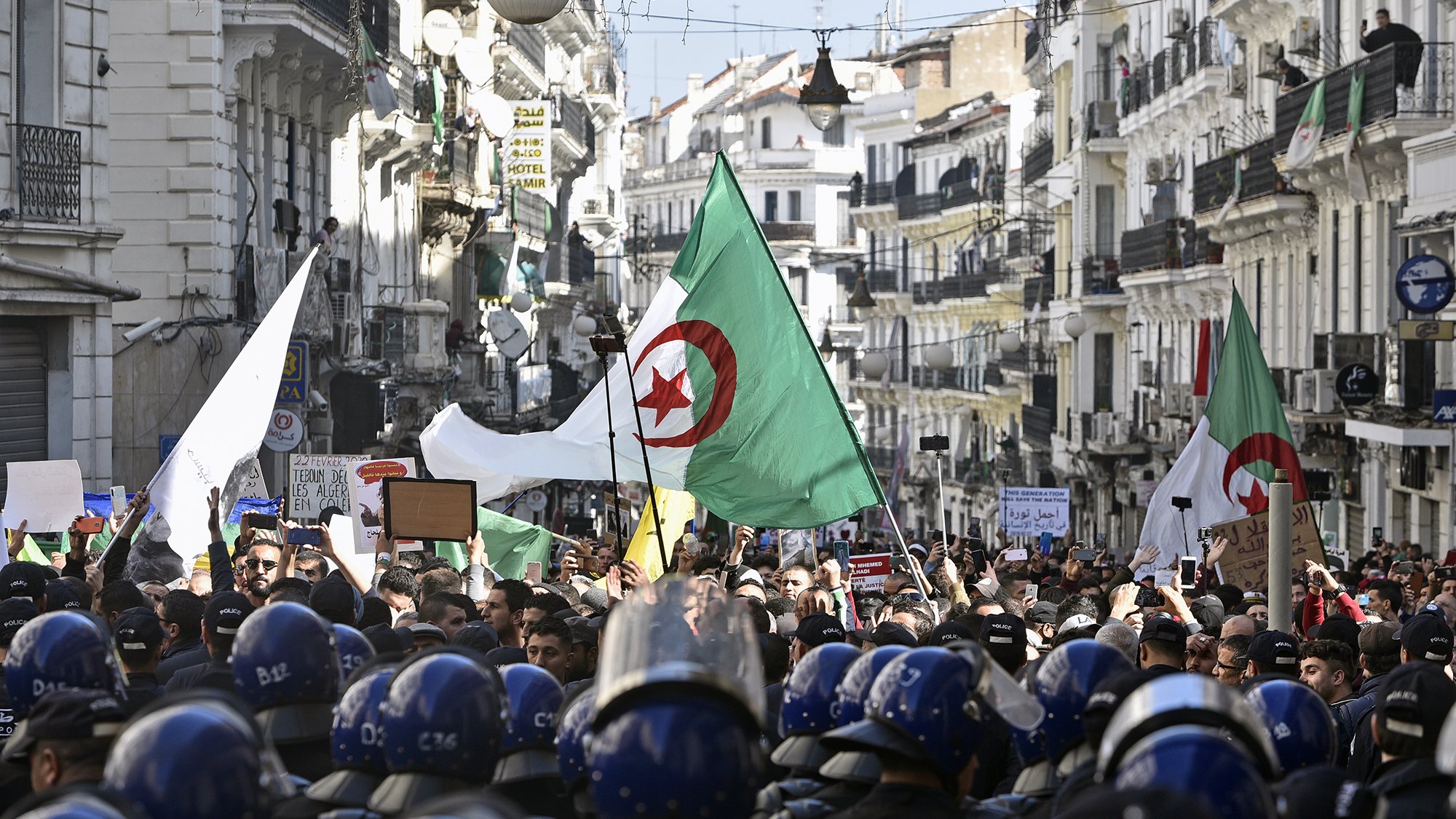 The image depicts a large protest scene in a street lined with buildings. Many people are gathered, holding flags, including Algerian flags, and signs. The crowd is diverse and appears to be demonstrating for a cause. In the foreground, a line of riot police in helmets is visible, standing in front of the protesters, indicating a tense atmosphere. The street is adorned with lights and decorations, suggesting it might be a significant location in the city. Overall, the image captures a moment of civic engagement and protest.