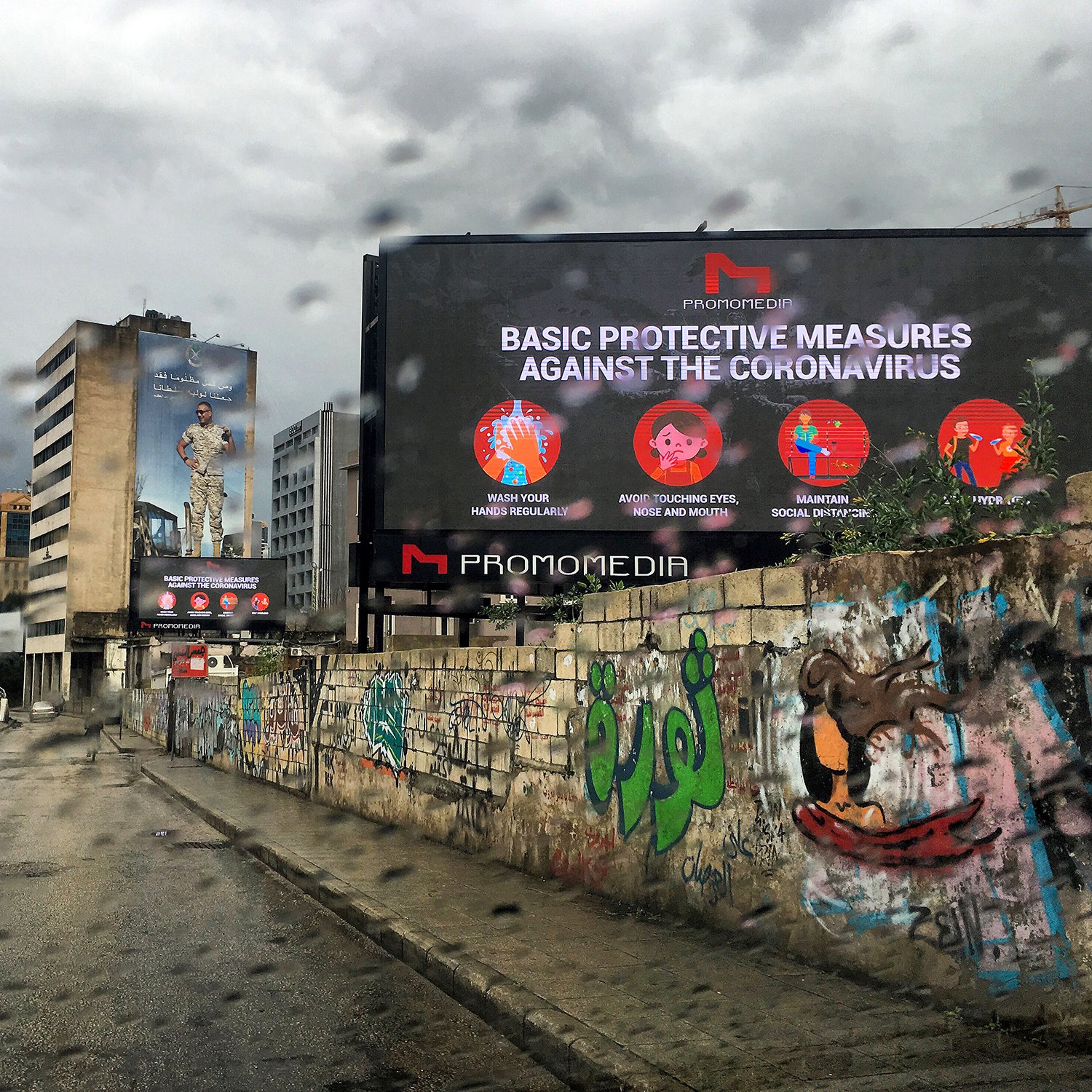 La imagen muestra una calle en un entorno urbano, con un ambiente lluvioso. Hay un letrero digital grande que presenta medidas de protección básicas contra el coronavirus. En el fondo, se pueden ver edificios y murales coloridos en las paredes, lo que añade un contraste visual a la escena. El cielo está nublado, lo que sugiere un clima gris y lluvioso.
