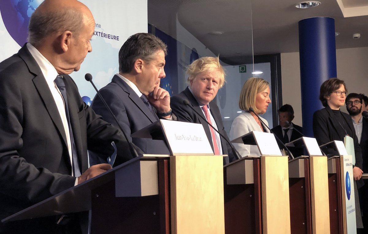 L'image montre une conférence de presse avec plusieurs personnes debout derrière des pupitres. Parmi eux, on peut voir des hommes en costume et une femme en tenue formelle. Ils semblent être en train de discuter ou d'écouter des questions, avec une atmosphère sérieuse. L'arrière-plan présente des éléments visuels représentant des thèmes internationaux, comme des logos ou des symboles géographiques.