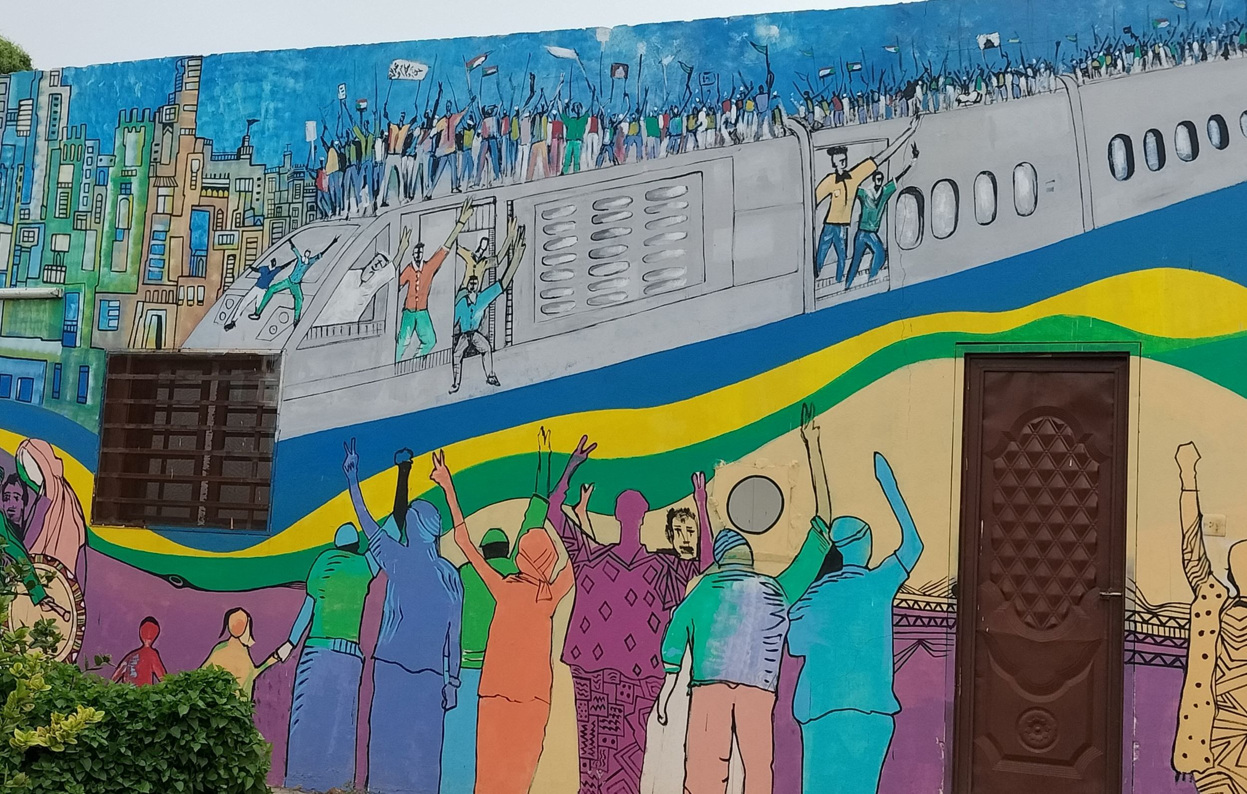 L'immagine rappresenta un murale colorato su un muro. La scena ritrae una folla di persone che sembra celebrare o protestare, con alcune figure che sollevano le braccia in segno di gioia o di richiesta. Sullo sfondo, c'è un aereo che si staglia sopra una città stilizzata, caratterizzata da edifici e una vivace palette di colori. I colori utilizzati nel murale sono brillanti e vivaci, riflettendo un senso di energia e movimento. La composizione comunica un forte messaggio di unità e di movimento collettivo.