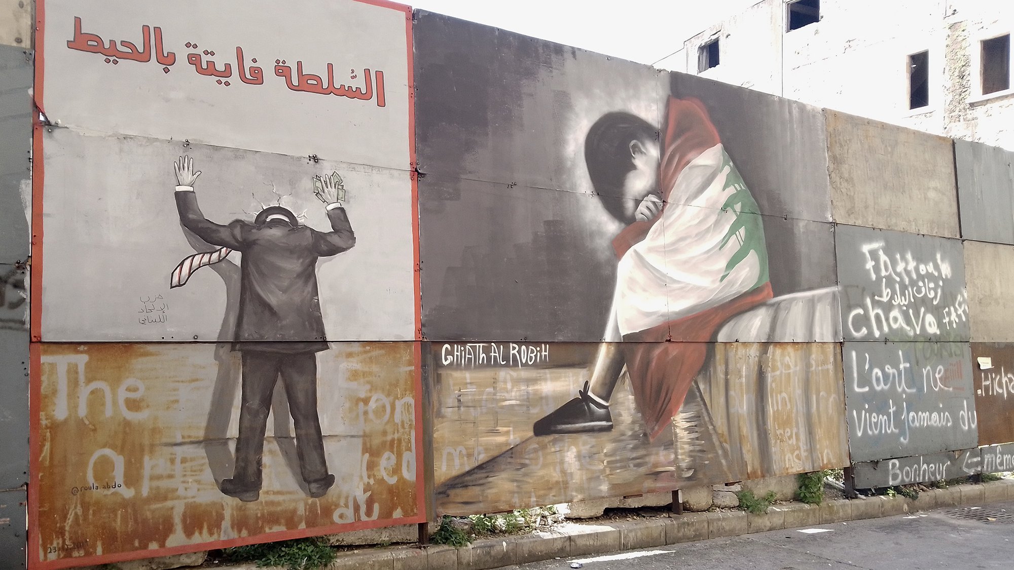 L'image montre un mur recouvert de plusieurs peintures murales. À gauche, on voit une figure humaine en costume, les mains levées, comme en signe de désespoir ou de soumission, avec une inscription en arabe qui pourrait évoquer des thèmes de répression. À droite, une autre figure, probablement d'une personne en train de pleurer ou de s'asseoir tristement, est représentée, vêtue d'une robe aux couleurs d'un drapeau, ce qui pourrait symboliser une forte connexion à une identité culturelle ou nationale. Le fond comporte également des mots ou des phrases en français et en arabe, ajoutant une dimension de commentaire social ou politique à l'œuvre. L'ensemble évoque des thèmes de lutte, d'identité et d'émotion.