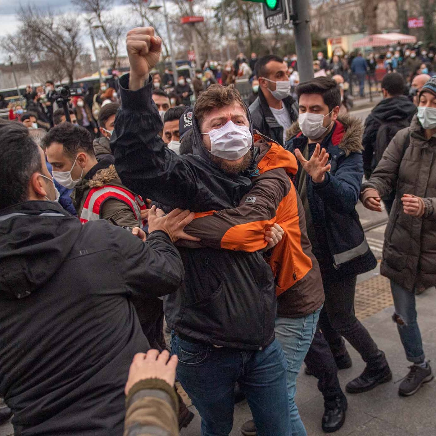The image depicts a scene of confrontation during a protest or demonstration. Several individuals are gathered, with some appearing to engage in a physical altercation. One person, wearing a black jacket and a mask, is raising a fist, indicating a strong emotional response. People around them seem to be trying to intervene or separate those involved in the conflict. The setting appears to be an urban area, likely near a traffic light, with a backdrop of buildings and other demonstrators. The atmosphere seems tense and charged.