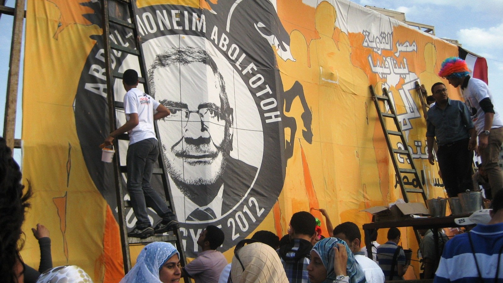 تظهر الصورة مجموعة من الأشخاص يعملون على رسم وإعداد لوحة ضخمة تحمل صورة وشعار أحد المرشحين الانتخابيين، حيث يقوم بعضهم بتلوين اللوحة باستخدام سلالم. الخلفية برتقالية مع تفاصيل أخرى، ويبدو أن الحدث مرتبط بالانتخابات في عام 2012. يظهر أفراد يرتدون ملابس متنوعة، بما في ذلك بعض الأشخاص الذين يرتدون أغطية رأس تحمل ألوان مختلفة. أجواء العمل والاجتهاد تسيطر على المشهد.