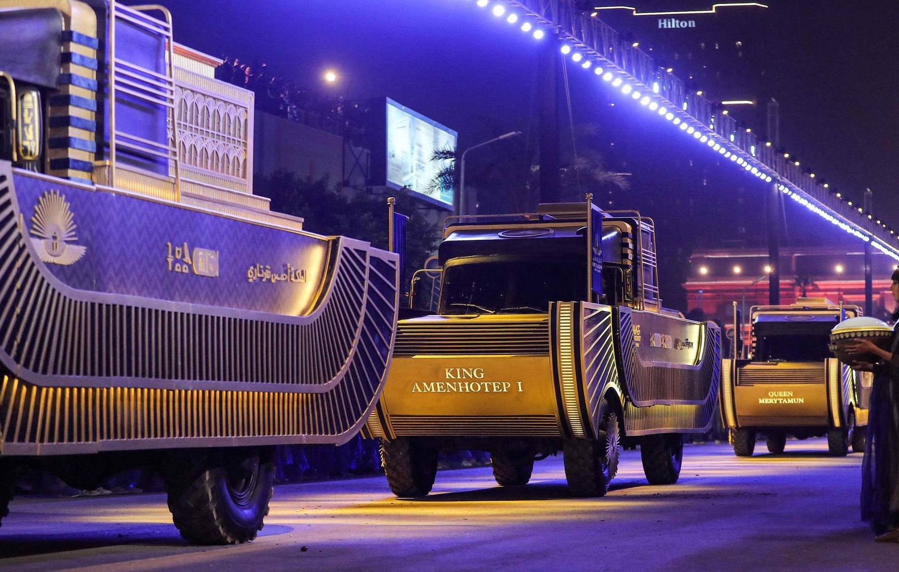 L'image montre un défilé nocturne avec des camions décorés, probablement pour une célébration ou un événement spécial. Les camions portent des inscriptions, dont "KING AMENHOTEP I", et sont éclairés par des lumières vives. En arrière-plan, on aperçoit un bâtiment, peut-être un hôtel, et des foules observant l'événement. Un personnage en costume traditionnel est également visible, ajoutant à l'atmosphère festive et culturelle.