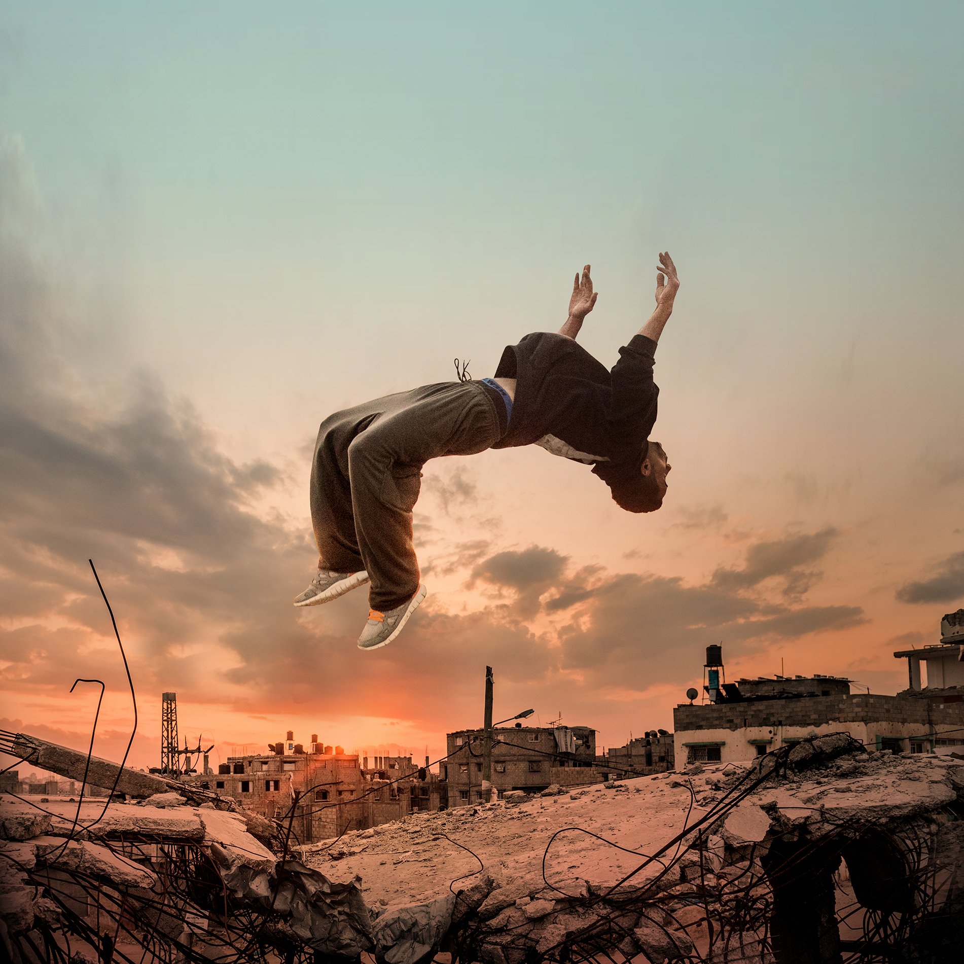 L'immagine mostra un giovane che sembra fluttuare o saltare sopra un paesaggio urbano distrutto, con scarti di costruzione e rovine. Sullo sfondo, si possono vedere nuvole e un cielo al tramonto, che creano un'atmosfera drammatica. La silhouette del ragazzo è evidenziata dalla luce, evidenziando un senso di libertà e movimento in un contesto desolato.