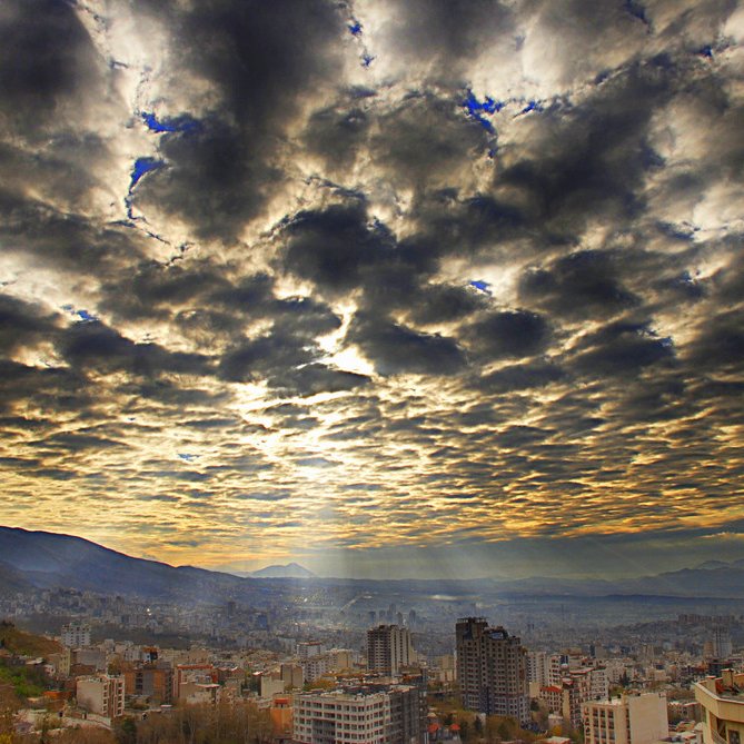 Cette image montre un paysage urbain avec des bâtiments s'étendant dans la vallée, sous un ciel dramatique rempli de nuages. Les nuages sont denses et variés, créant une texture visuelle intéressante, tandis que des rayons de lumière percent à travers eux, illuminant partiellement la ville en contrebas. On peut également apercevoir des montagnes à l'arrière-plan, ajoutant une dimension naturelle à la scène. L'atmosphère est à la fois majestueuse et sereine, capturant un moment poétique de la journée.