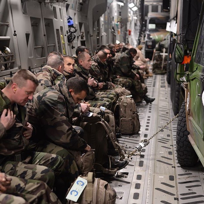 L'image montre un groupe de soldats assis à l'intérieur d'un véhicule militaire ou d'un avion de transport. Ils portent des uniformes camouflage et sont équipés de sacs à dos. Certains semblent attentifs et concentrés, tandis que d'autres sont plus détendus. Le cadre est sombre et fonctionnel, typique des espaces militaires, avec des sièges alignés sur les côtés et un véhicule militaire garé à l'intérieur.