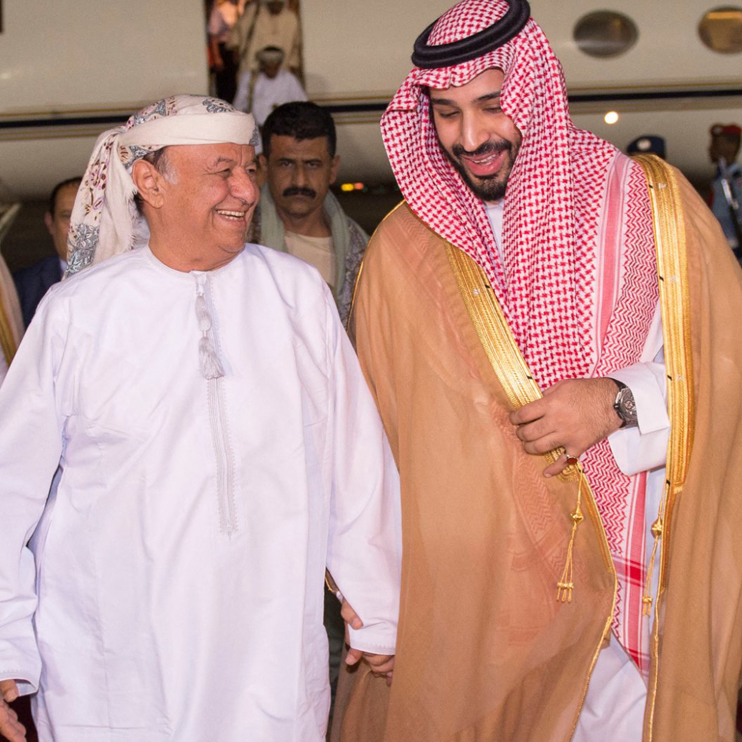 La imagen muestra a dos hombres sonriendo y caminando juntos. Uno de ellos lleva un traje tradicional árabe, con una túnica blanca y un tocado, mientras que el otro viste un manto largo con adornos y una kufiya. Ambos parecen estar en un ambiente festivo, posiblemente en el aeródromo, y hay un avión privado visible al fondo. La expresión de ambos es amistosa y cordial.
