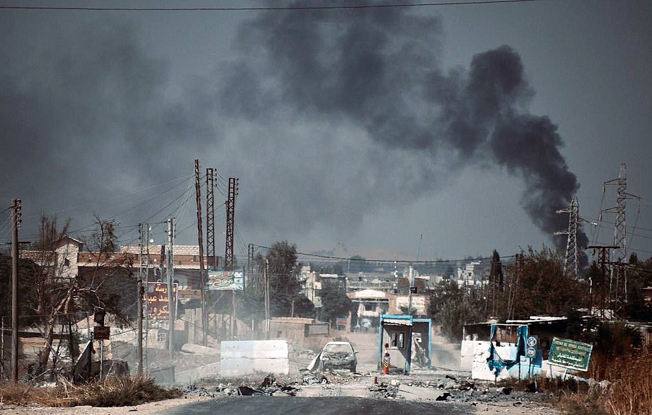 The image depicts a desolate street under a dark, smoke-filled sky. In the background, billowing plumes of black smoke suggest an ongoing fire or explosion. The foreground shows a damaged road with debris, potentially hinting at a conflict zone. There are signs of destruction, including burnt vehicles and abandoned structures, contributing to an overall atmosphere of turmoil and devastation. The landscape appears barren with a few power lines and distant buildings, reflecting the impact of violence on the area.