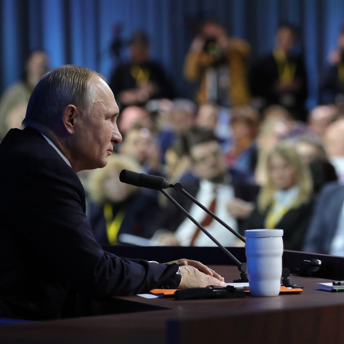 L'image montre un homme assis à une table de conférence, parlant peut-être lors d'une conférence de presse ou d'un discours. Il semble concentré et fait face à un public, qui est partiellement visible en arrière-plan. On aperçoit des journalistes prenant des notes ou photographiant. Des micros sont placés devant lui, et une tasse blanche est posée sur la table. L'atmosphère paraît sérieuse et attentive.