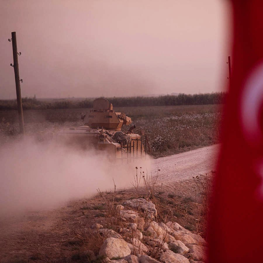 در این تصویر، یک تانک نظامی در حال حرکت در یک جاده خاکی به سمت جلو دیده می‌شود. در پس‌زمینه، میدان‌های سبز و طبیعتی آرام قرار دارد. در سمت چپ تصویر، پرچم ترکیه با نشانه‌های مشخص آن قرار دارد. غبار و دود ناشی از حرکت تانک فضای تصویر را پوشانده و حس تحرک و فعالیت نظامی را منتقل می‌کند. نور ملایم غروب یا صبح نیز به فضای کل تصویر جذابیت بیشتری بخشیده است.