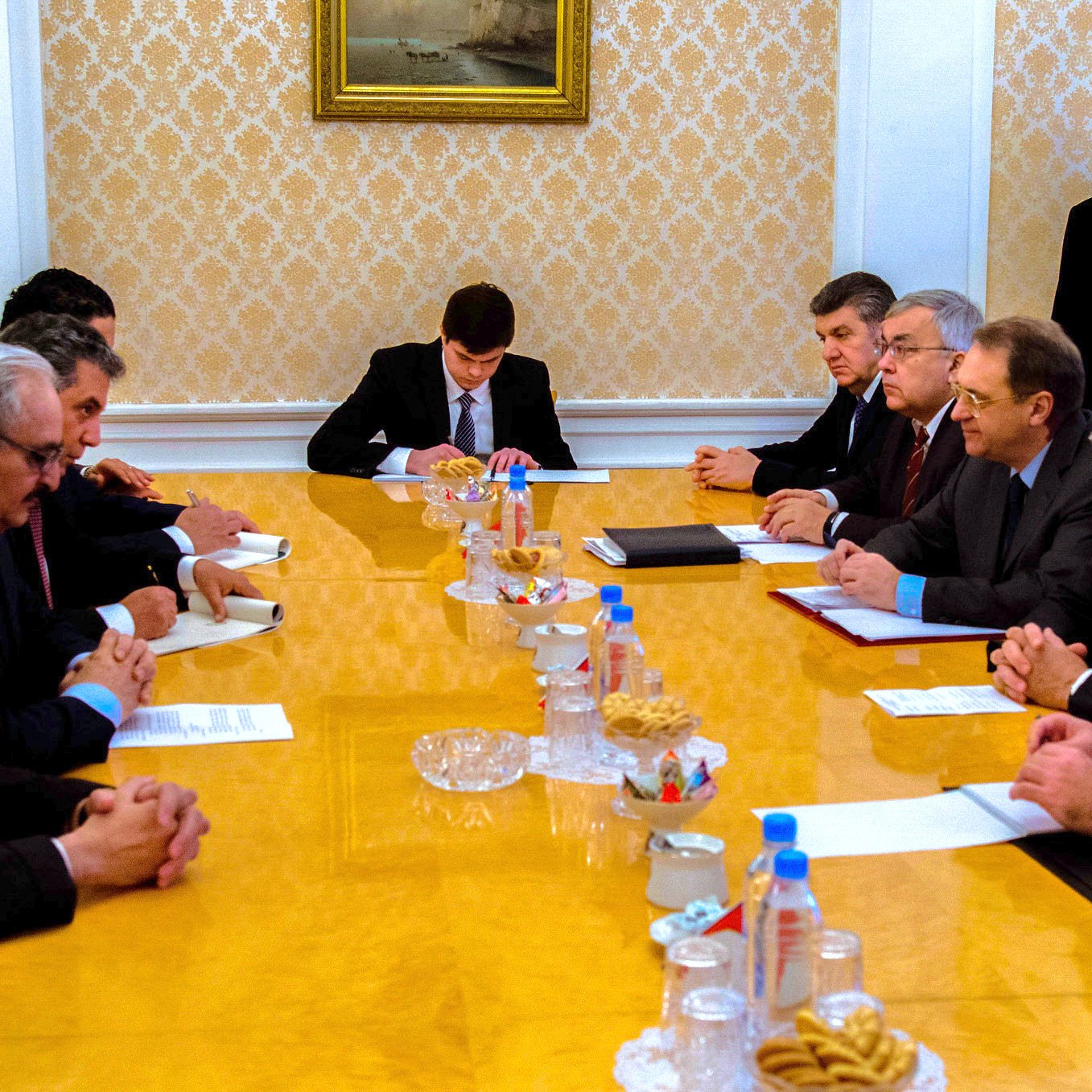 L'image présente une réunion autour d'une grande table ovale en bois. Plusieurs hommes sont assis des deux côtés, en tenue formelle, semblant être en discussion ou en train de négocier. Au centre de la table, il y a des documents et des canapés, ainsi qu'une assiette de collations. En arrière-plan, on peut voir des personnes observant la réunion, ce qui indique que c'est un moment important. L'atmosphère semble sérieuse et concentrée.