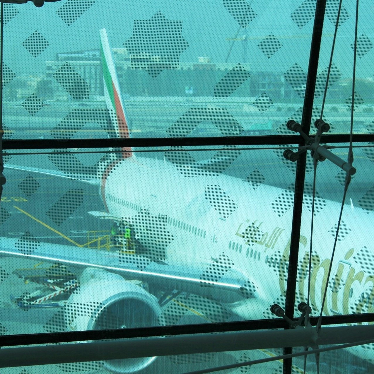 La imagen muestra un avión de la aerolínea Emirates estacionado en un aeropuerto. Se puede ver parte del fuselaje y el ala del avión a través de un gran ventanal con un diseño en las ventanas. Hay trabajadores en la plataforma que parecen estar realizando tareas de mantenimiento o preparación, y se puede observar un entorno aeroportuario con otros edificios y maquinaria en el fondo. La atmósfera es luminosa, aunque un poco difusa debido al cristal.