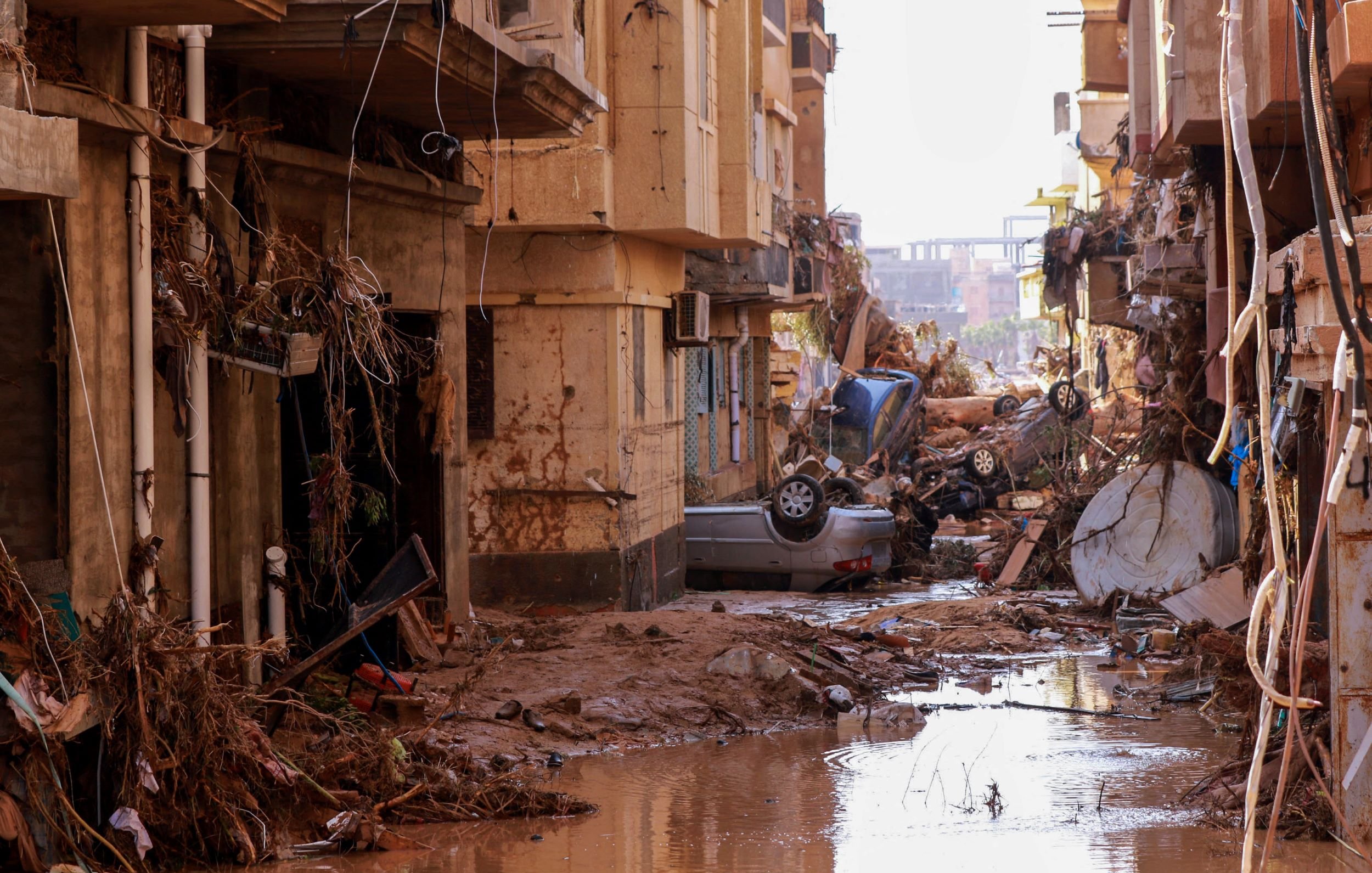 الصورة تظهر مشهداً من منطقة تعرضت لأضرار جسيمة نتيجة كارثة طبيعية، ربما تكون فيضانات أو عواصف. تظهر في الصورة مباني متضررة وجدران متهدمة، وهناك حطام متناثر في الشوارع. كما تُظهر الصورة سيارات مقلوبة أو متضررة، ومياه راكدة تجمعت في الشوارع. المشهد يعكس الأثر المدمر الذي أحدثته الكارثة، مما يدلل على حالة الفوضى والمأساة التي خلفتها.
