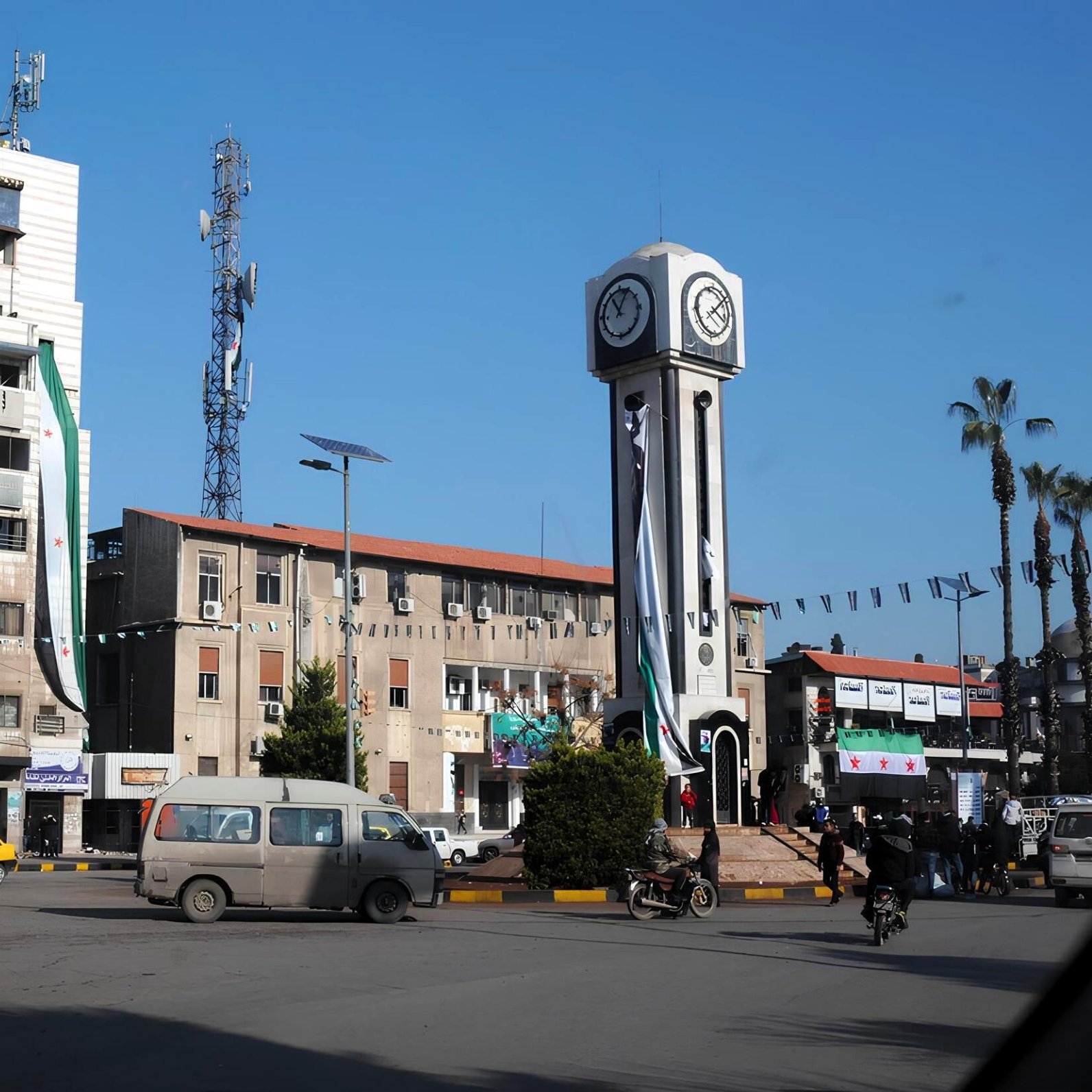 Una piazza con un grande orologio, edifici e alcuni veicoli in movimento.