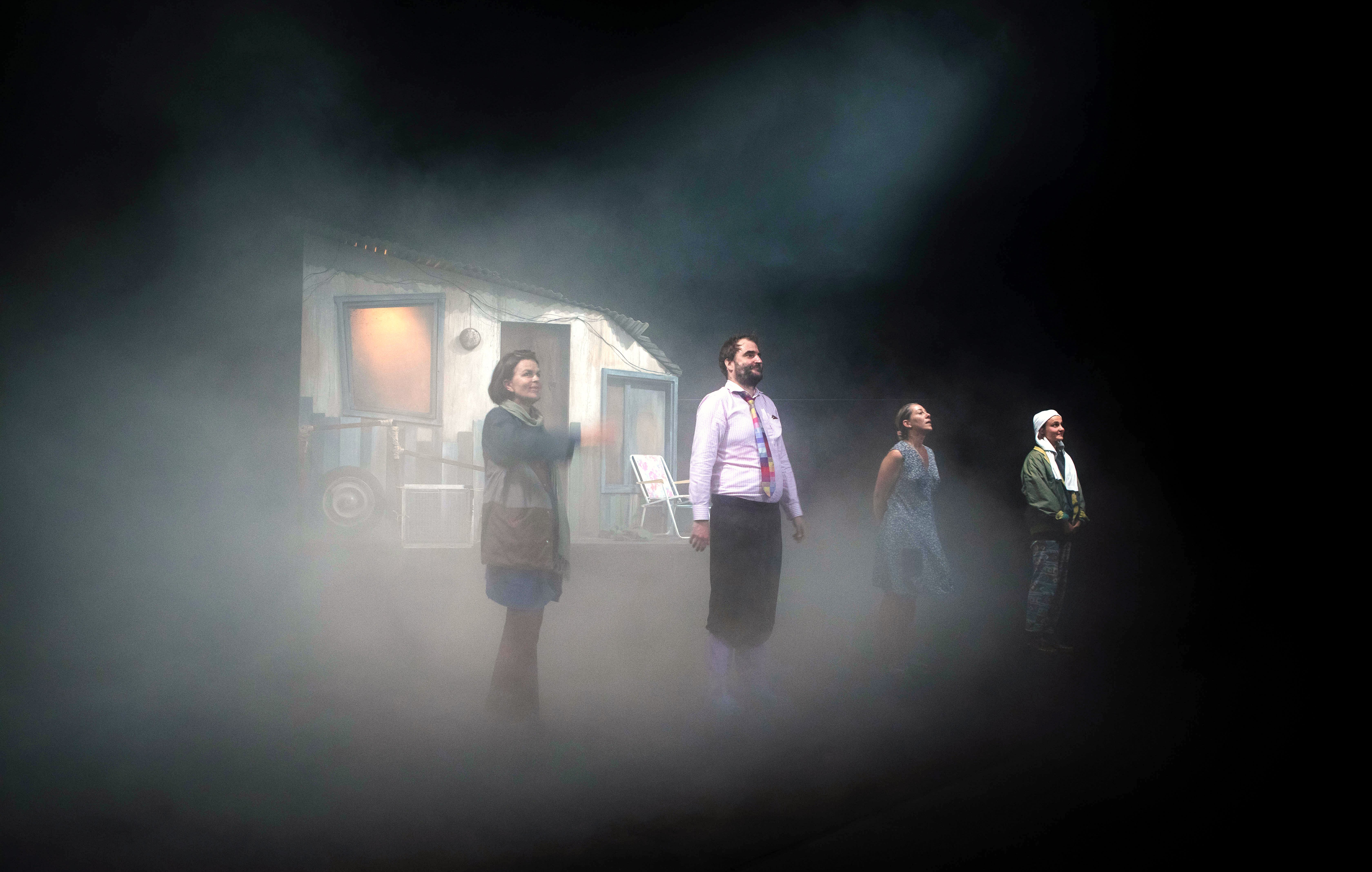 L'image présente une scène de théâtre avec quatre personnages en position debout sur scène. Ils se trouvent dans une atmosphère brumeuse, ce qui ajoute une ambiance mystérieuse. En arrière-plan, on peut apercevoir une petite maison ou un décor qui semble inhabitée. Les personnages sont vêtus de manières diverses, ce qui suggère des rôles ou des traits de caractère différents. La lumière semble tamisée, renforçant l'effet dramatique de la scène.
