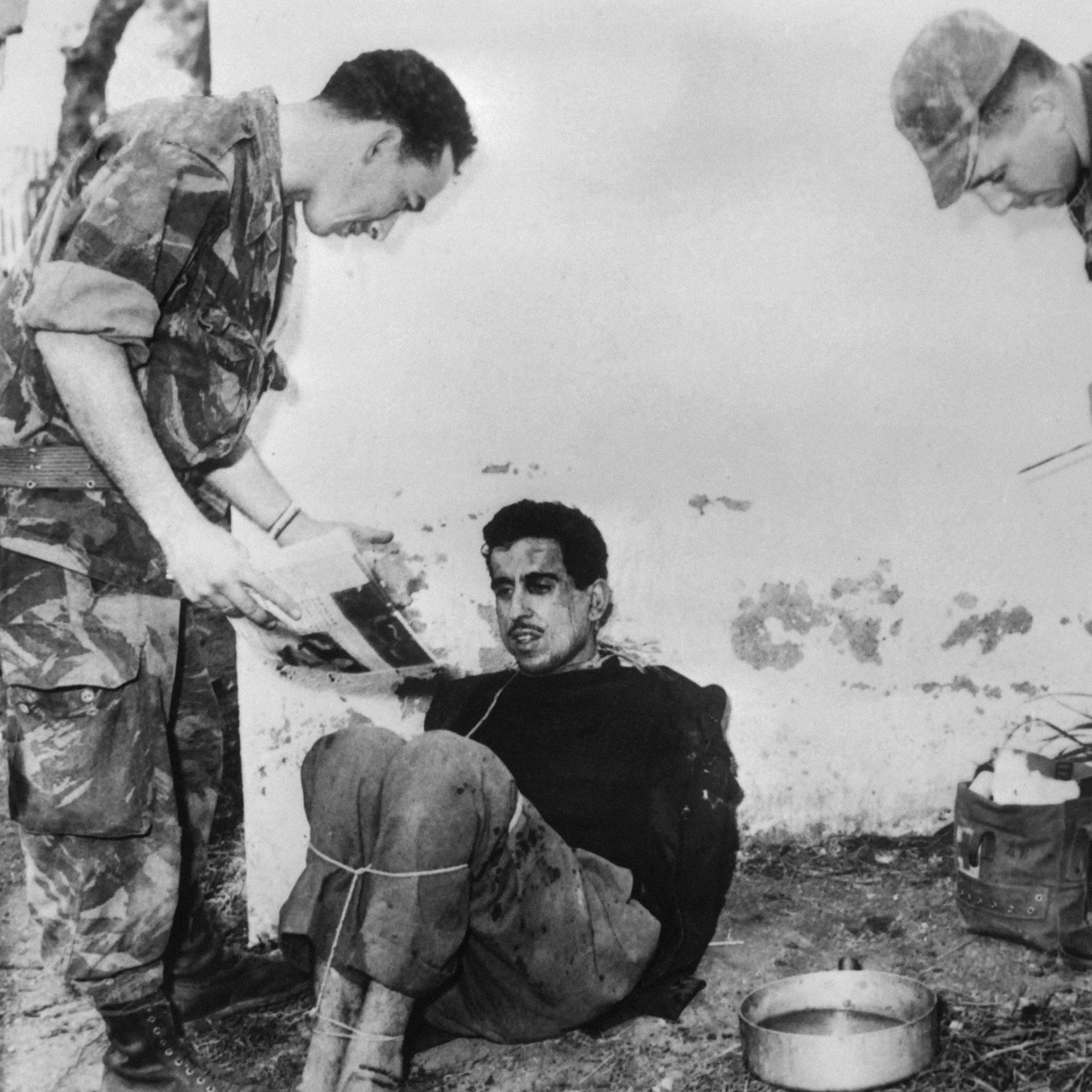 الصورة تظهر مجموعة من الجنود أثناء استجواب رجل يبدو أنه محتجز. الرجل جالس على الأرض ومربوط. الجنود يظهرون بزيهم العسكري، ويبدو أنهم يركزون على الوثائق أو الصور. البيئة المحيطة تبدو خارجية وملوثة بعض الشيء، مما يوحي بمكان غير رسمي. تعكس الصورة أجواء توتر وصعوبة.