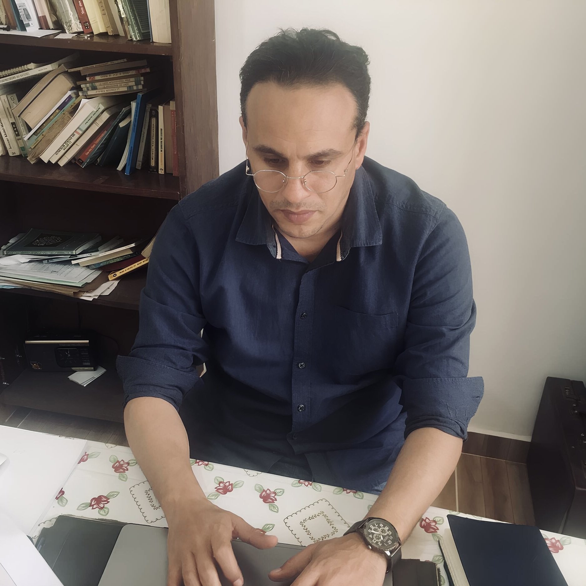 Nell'immagine si vede un uomo seduto a una scrivania. Indossa una camicia blu e occhiali. È concentrato mentre lavora su un computer portatile. La scrivania è disordinata, con fogli, un mouse e altri oggetti sparsi. Sullo sfondo si intravede una libreria con libri. L'ambiente sembra luminoso e semplice.