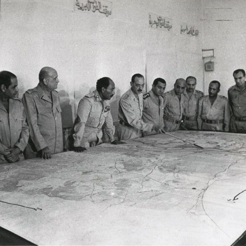 تظهر الصورة مجموعة من الرجال يرتدون زيًا عسكريًا، يجتمعون حول طاولة كبيرة تعرض خريطة. يبدو أن الاجتماع يدور حول التخطيط أو مناقشة استراتيجية ما. تعكس تعابير وجوههم التركيز والاهتمام بما يُناقش. الجدران خلفهم تبدو بسيطة، مع بعض الكتابات باللغة العربية. تُعطي الصورة انطباعًا عن موقف رسمي وعسكري.