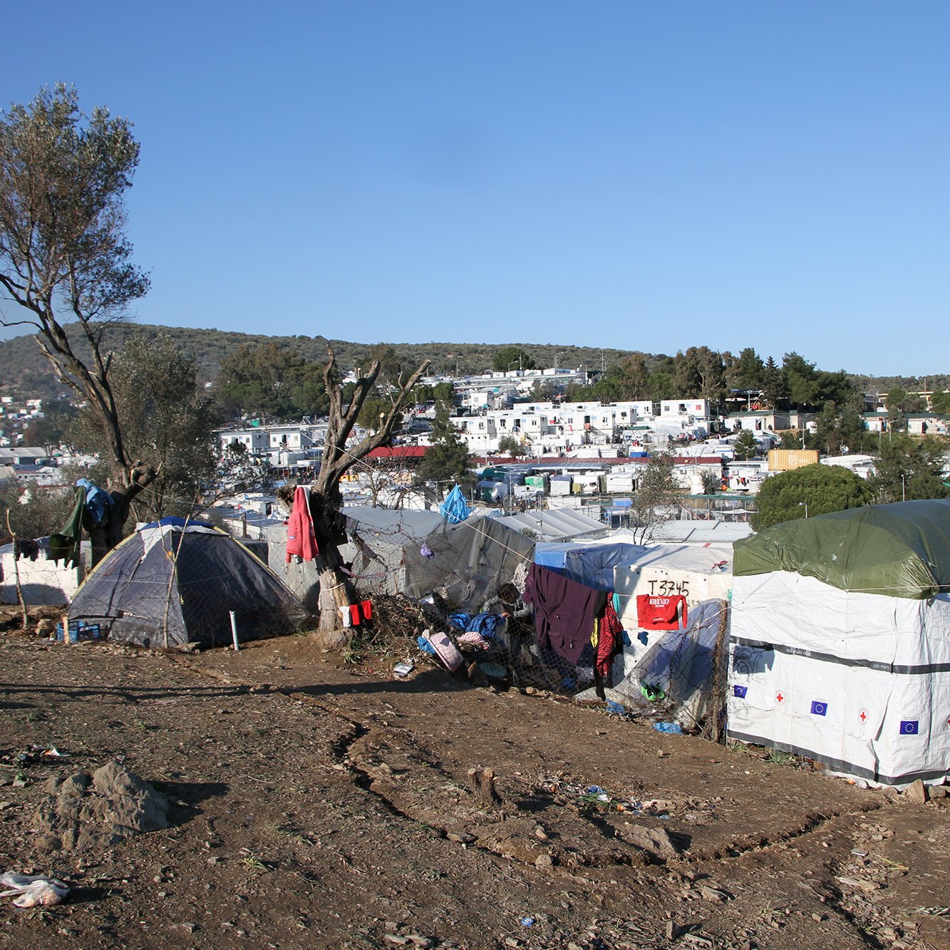 The image depicts a makeshift settlement in a rural or semi-urban area. Numerous tents are arranged on a slope, suggesting a temporary living situation. The landscape features sparse vegetation and a few trees, while in the background, there are more established buildings, possibly residences or facilities. The scene conveys a sense of hardship, with signs of disarray and poverty surrounding the tents. The sky is clear with little cloud cover, indicating a sunny day. A person stands in the foreground, observing the surroundings.