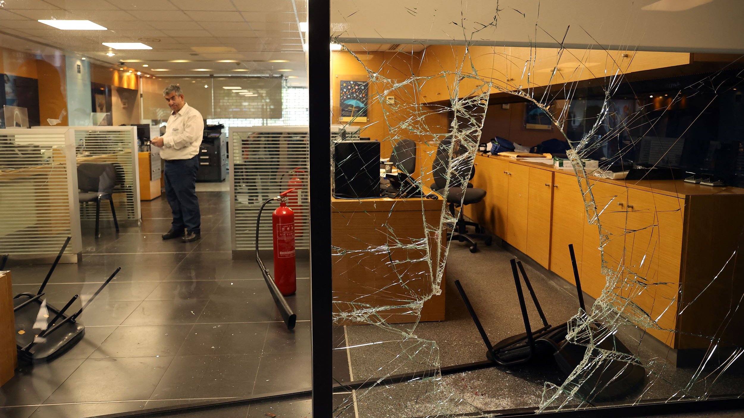 L'image montre un bureau avec des dégâts visibles. On peut voir une vitre brisée à l'avant, avec des fissures qui s'étendent sur toute sa surface. À l'intérieur, un homme est debout, semblant examiner la situation, tandis que des chaises sont renversées sur le sol. L'ambiance générale suggère qu'il y a eu un incident perturbateur dans cet espace de travail.