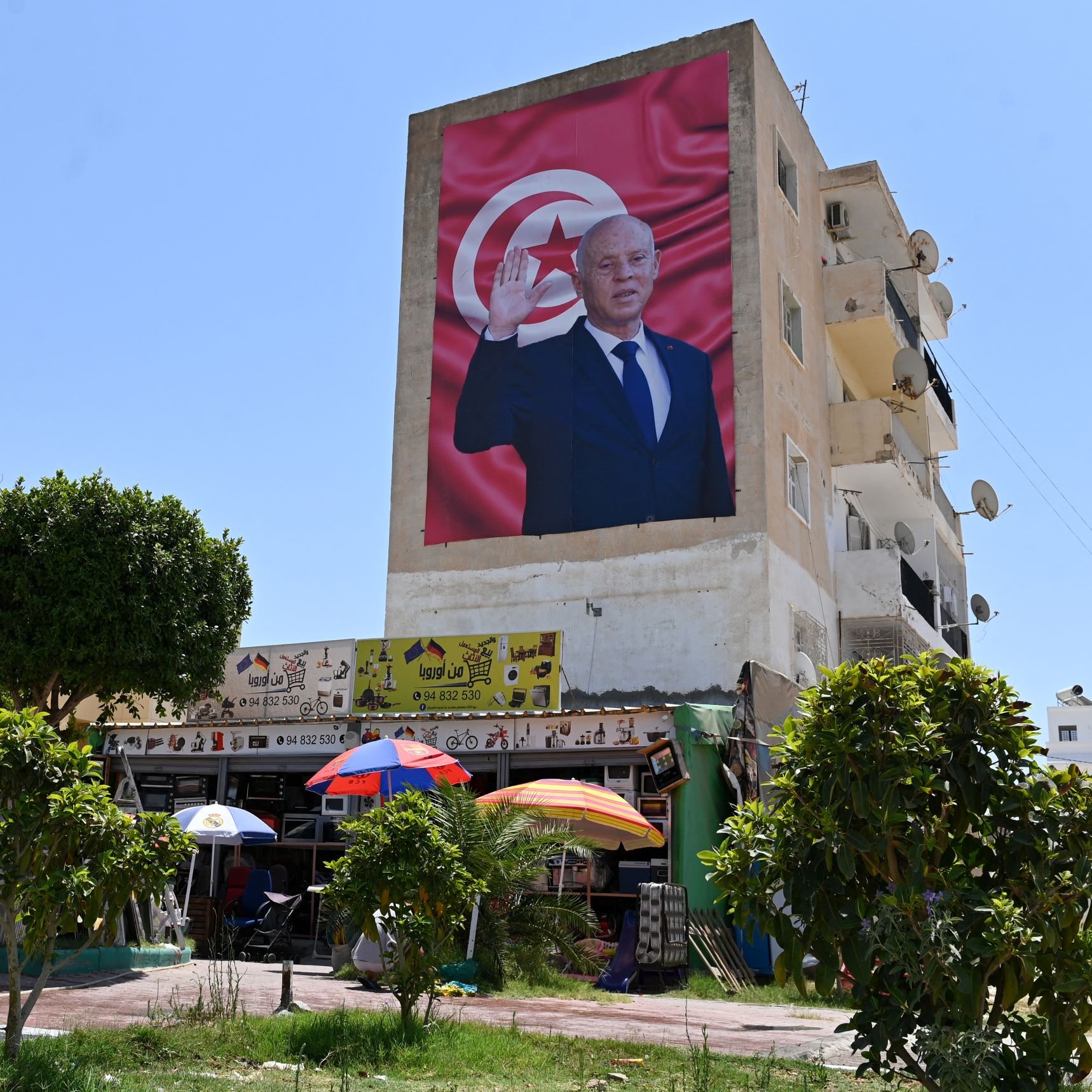 The image features a large mural on the side of a building, depicting a figure in formal attire, raising a hand in a gesture of greeting or salute. The background of the mural includes a red backdrop with a prominent depiction of the national flag of Tunisia. Below the mural, there is a scene of a small open area with umbrellas, likely providing shade for people sitting or gathering. Surrounding the area are some trees and bicycles, suggesting a lively community space. The sky is clear and blue, indicating a bright day.